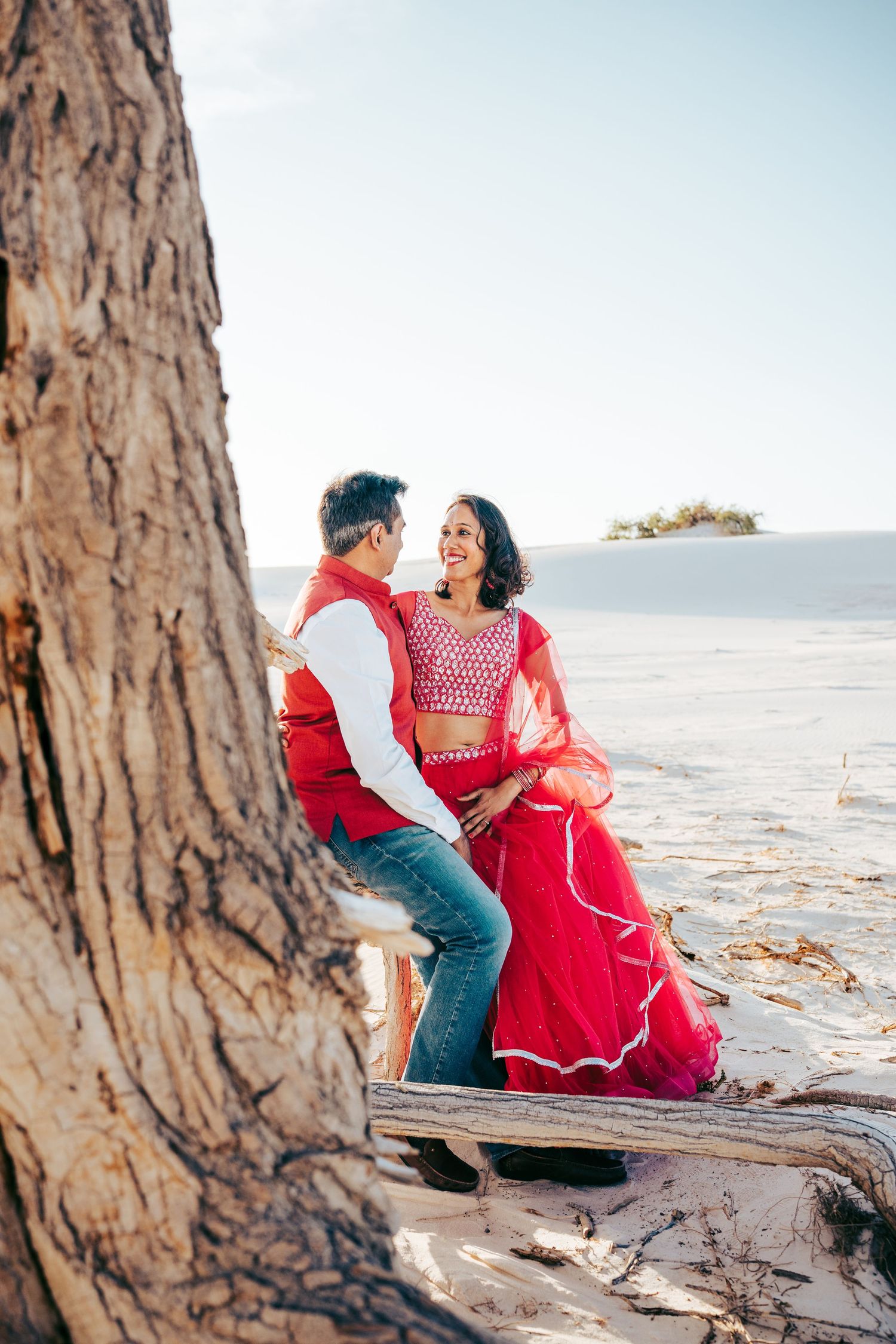 Smiling Indian couple in traditional red outfits sitting on driftwood framed by a large tree.
