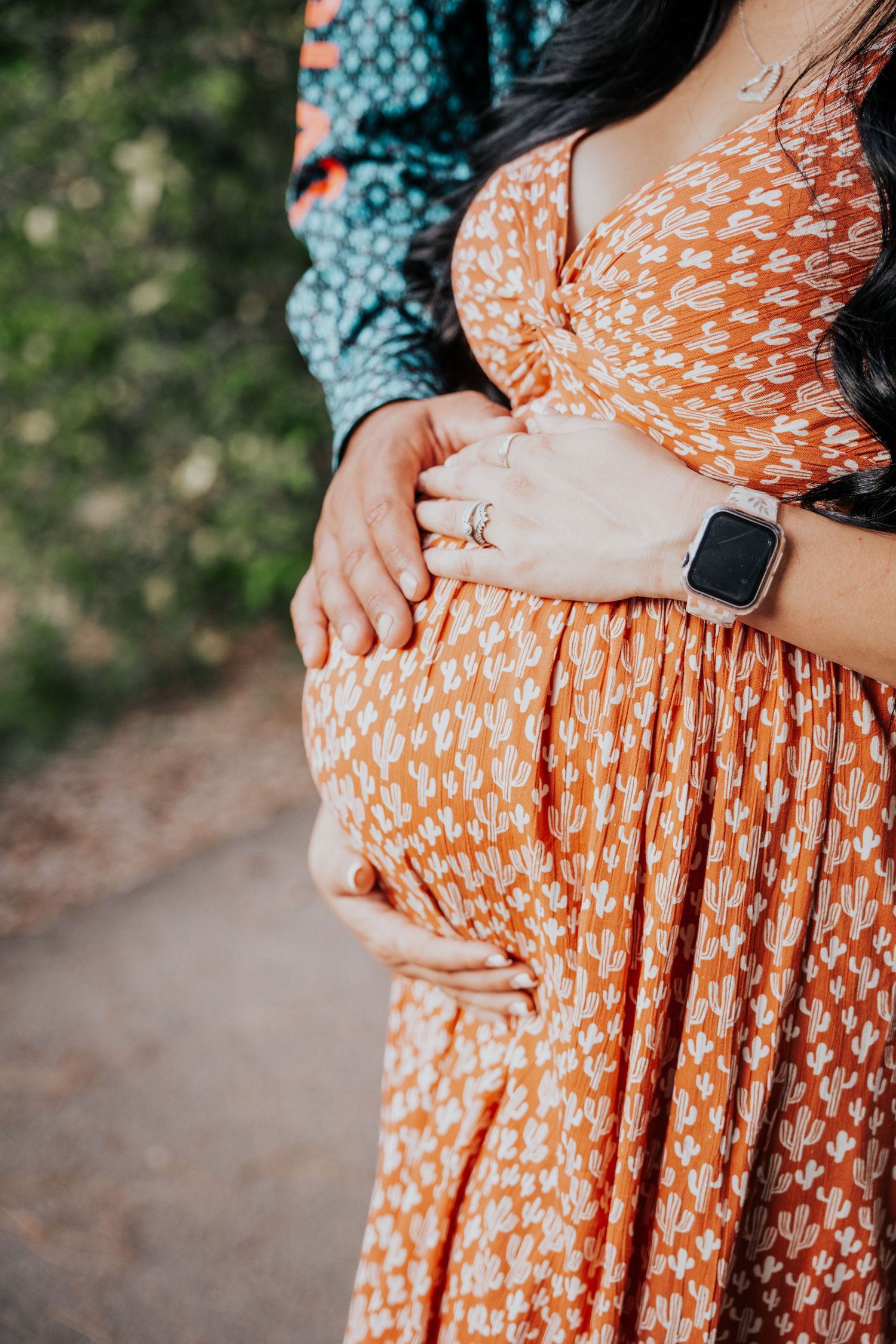 Close-up of pregnant belly with couples hands cradling bump, woman wearing orange floral dress and smartwatch.