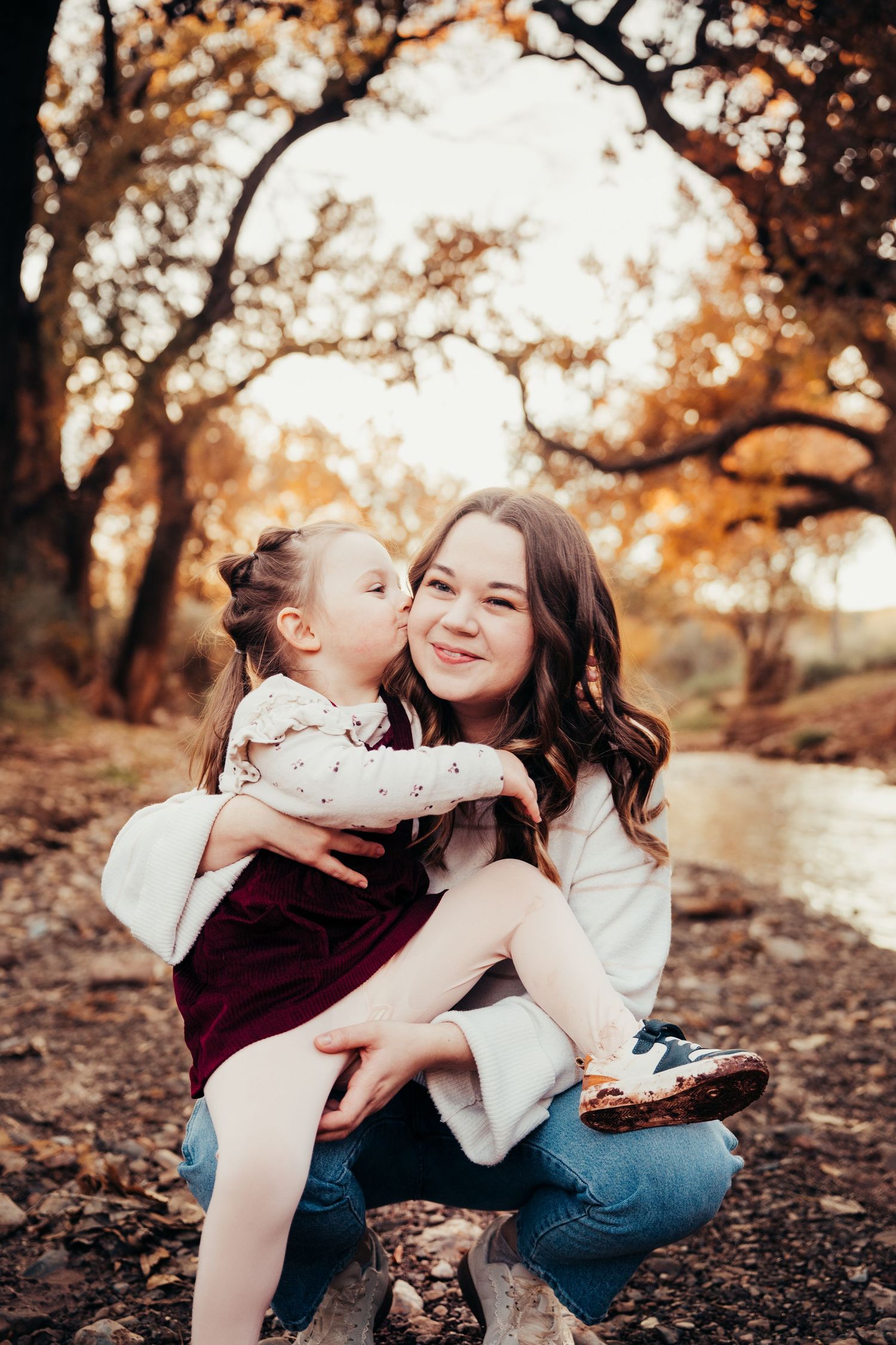 Sweet moment of toddler girl in burgundy dress kissing smiling mother on cheek in autumn park setting.