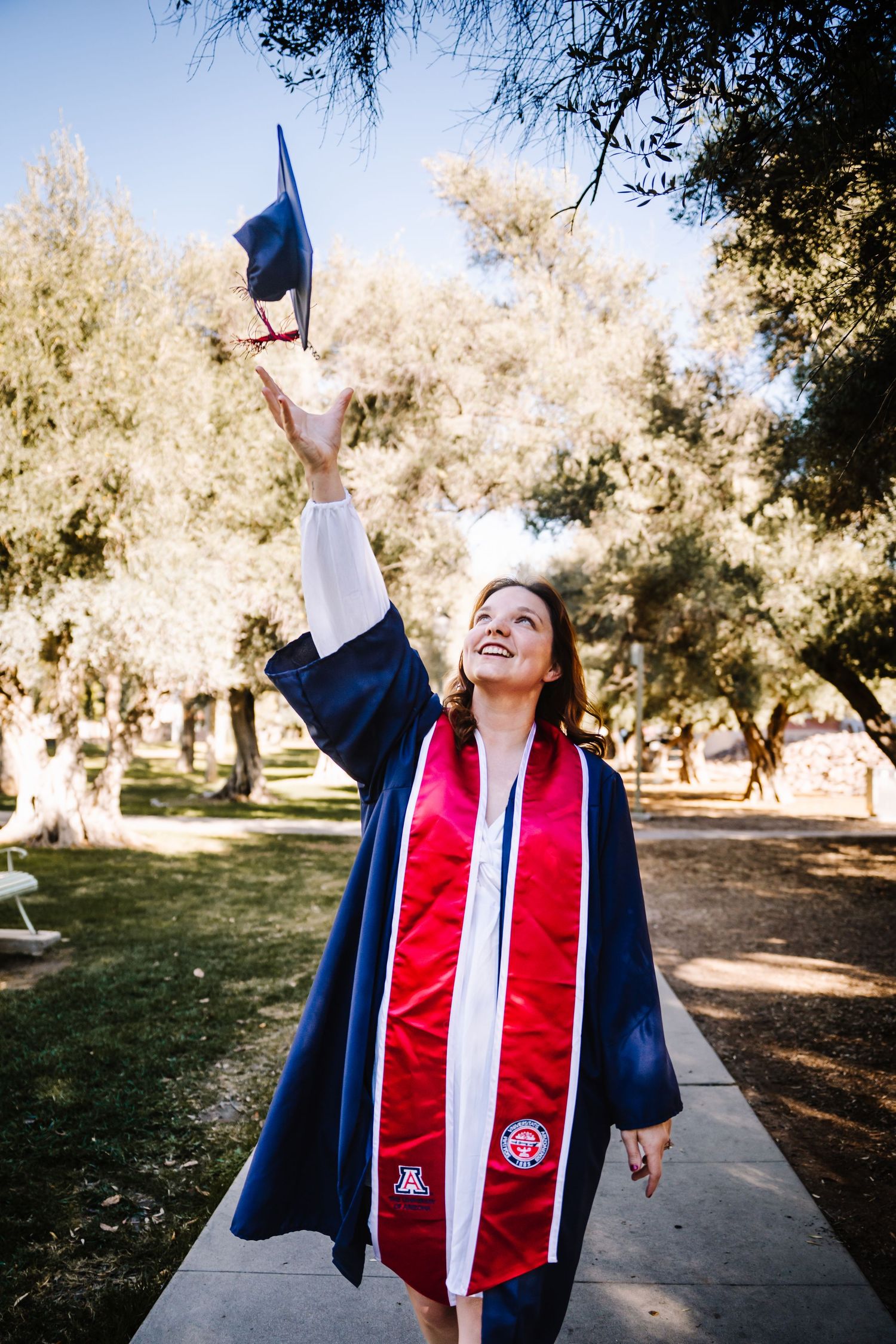 Graduate in navy blue gown tosses cap in the air outdoors, wearing red University of Arizona stole.