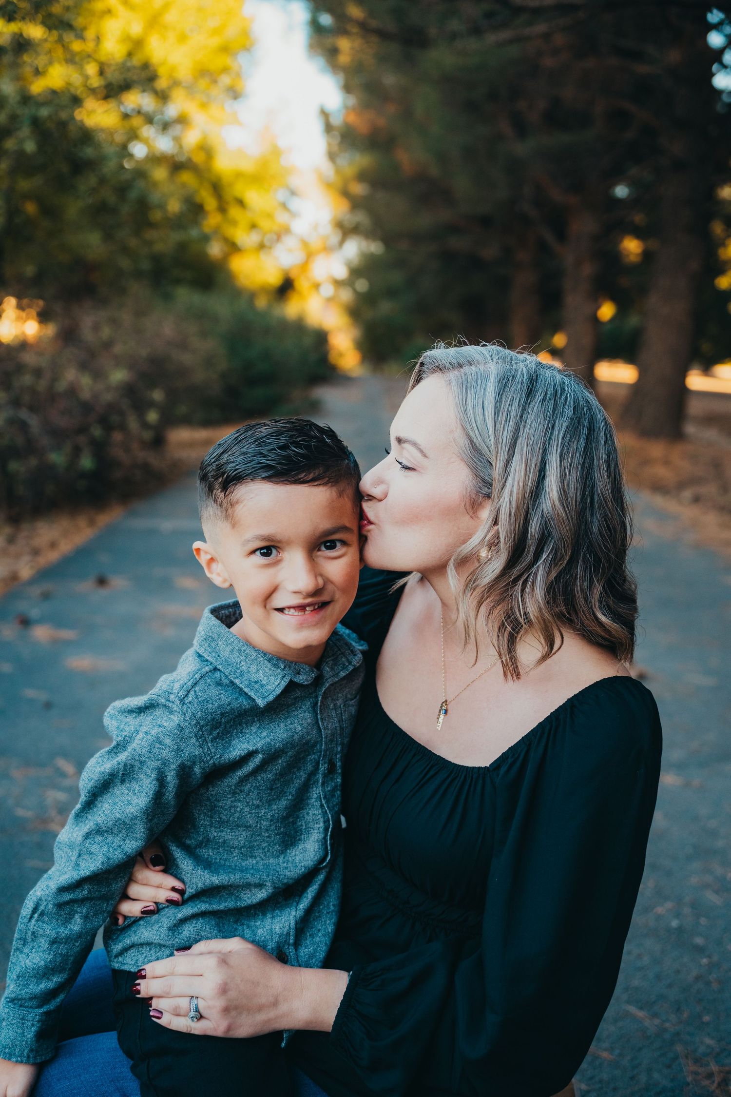 A smiling mother kissing her young son on the cheek while sitting outdoors in a sunlit park pathway.