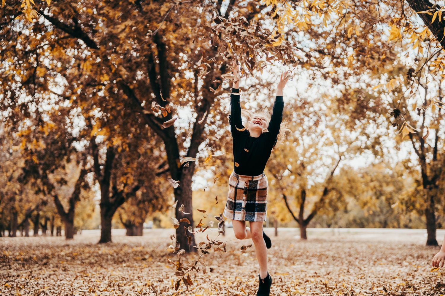 Young girl in black top and plaid skirt jumping and reaching for tree branches in autumn park.