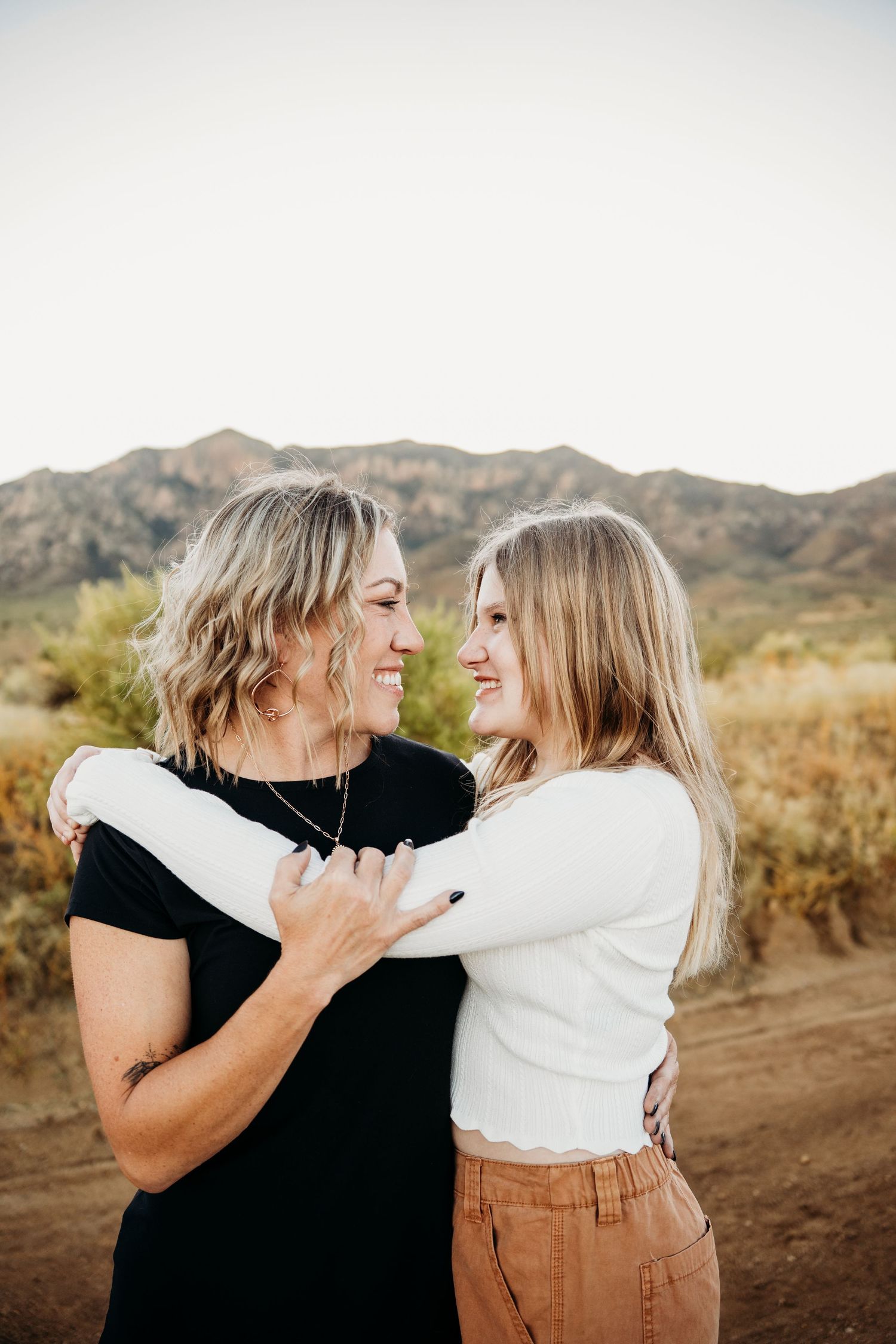 Two blonde women smiling and embracing on a desert trail with mountains behind them.