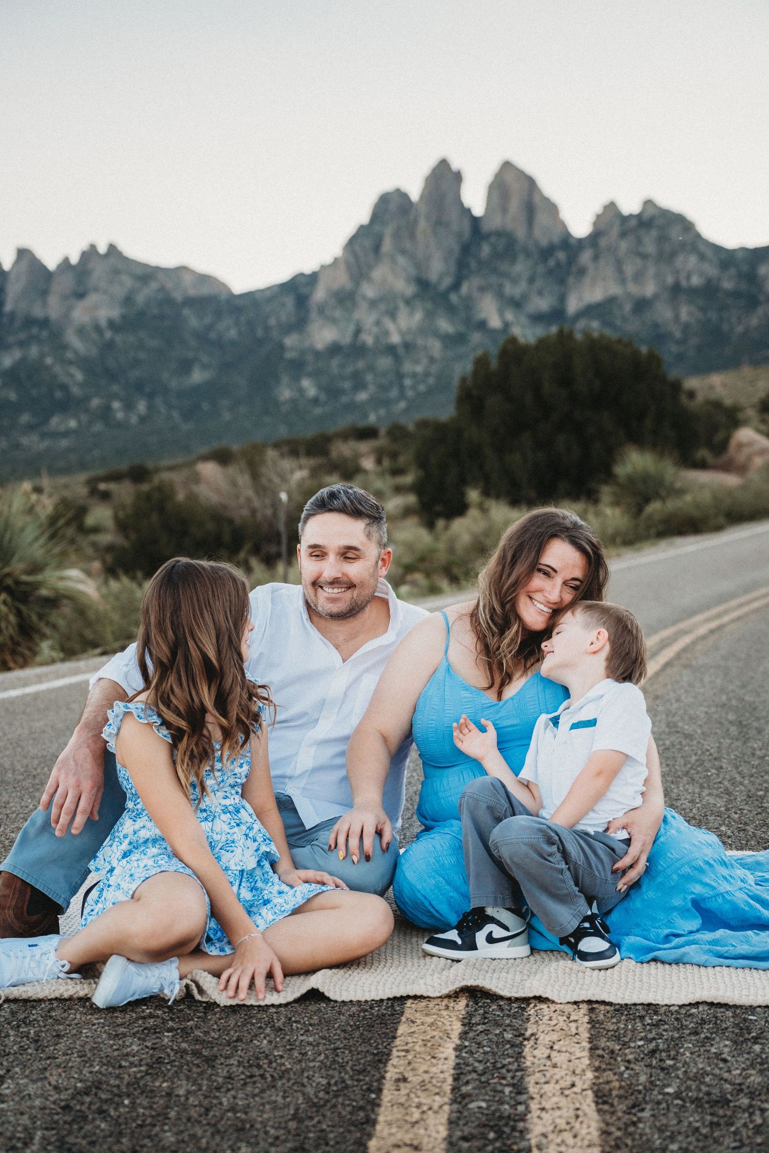 Joyful family of four seated on a road, mom and son sharing a sweet moment, mountains in background.