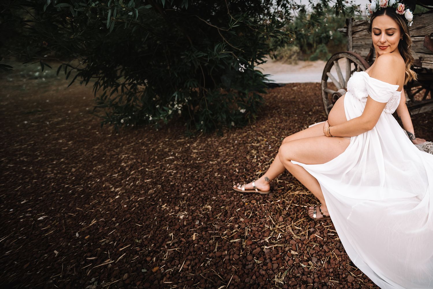 Elegant pregnant woman in white off-shoulder gown sitting gracefully outdoors near lush greenery.