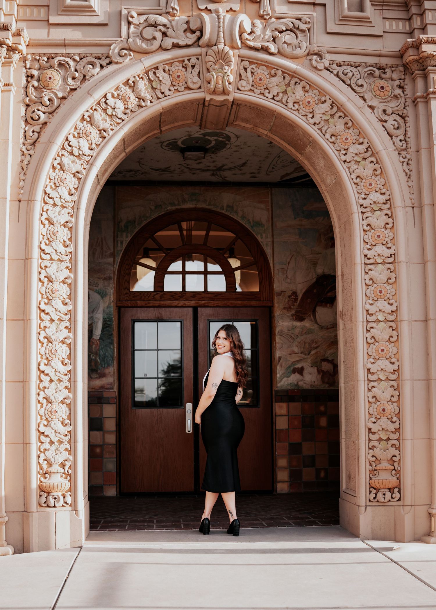 Woman in black fitted dress standing in elaborate Spanish Colonial Revival architectural archway.