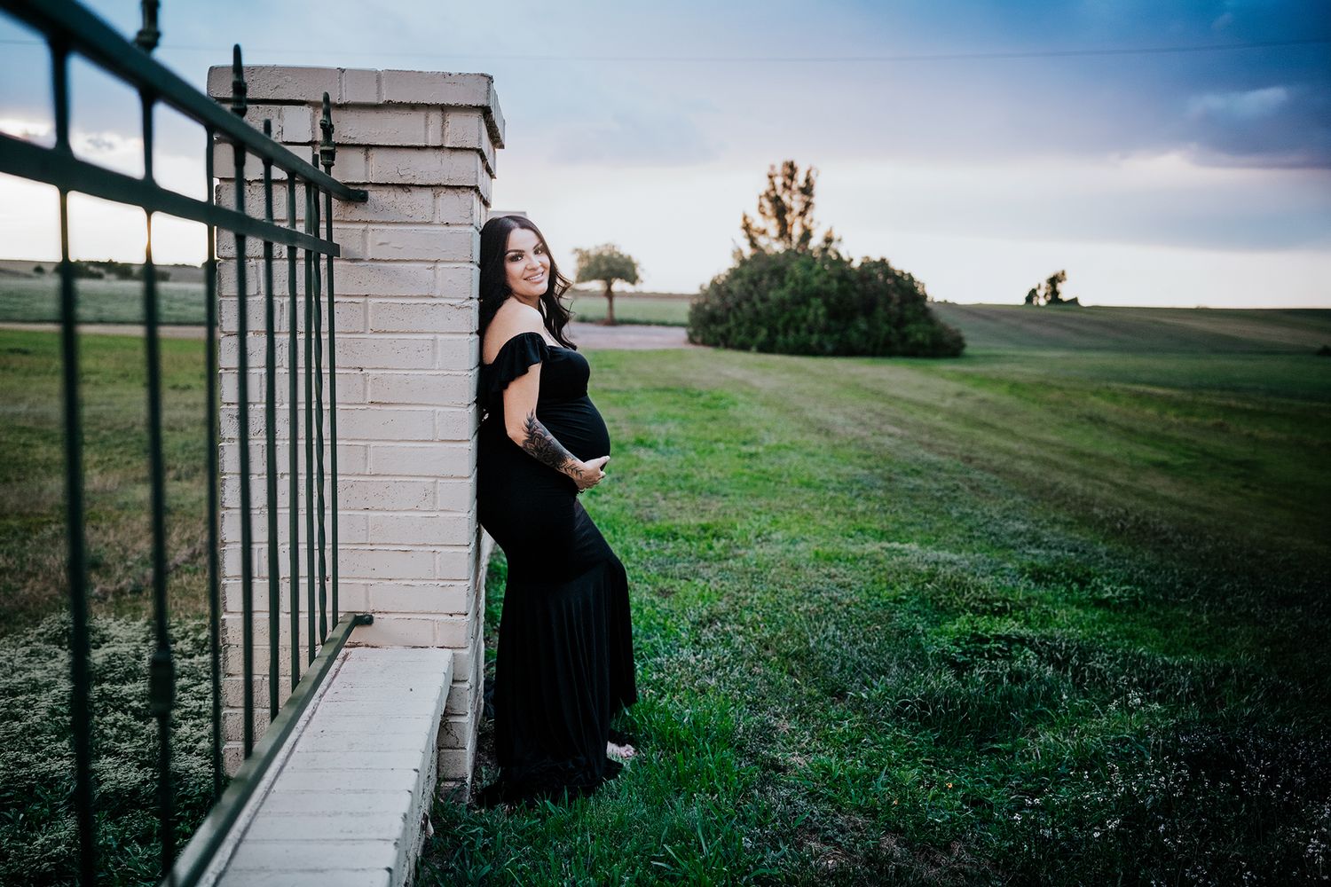 Pregnant woman in black gown leaning against brick wall in green field at dusk.