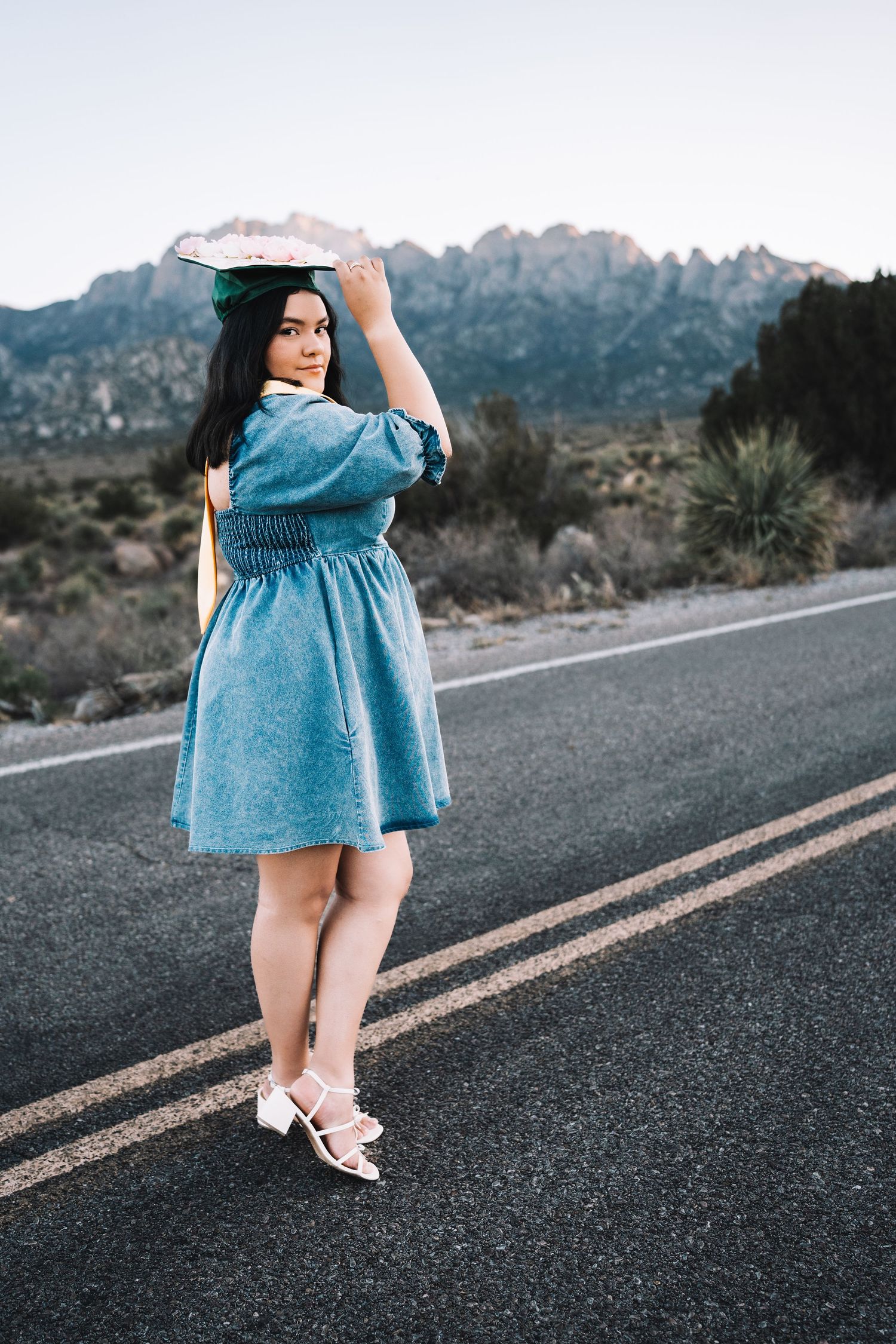 Graduate in a denim dress holding her decorated cap on a desert road with dramatic mountain scenery.