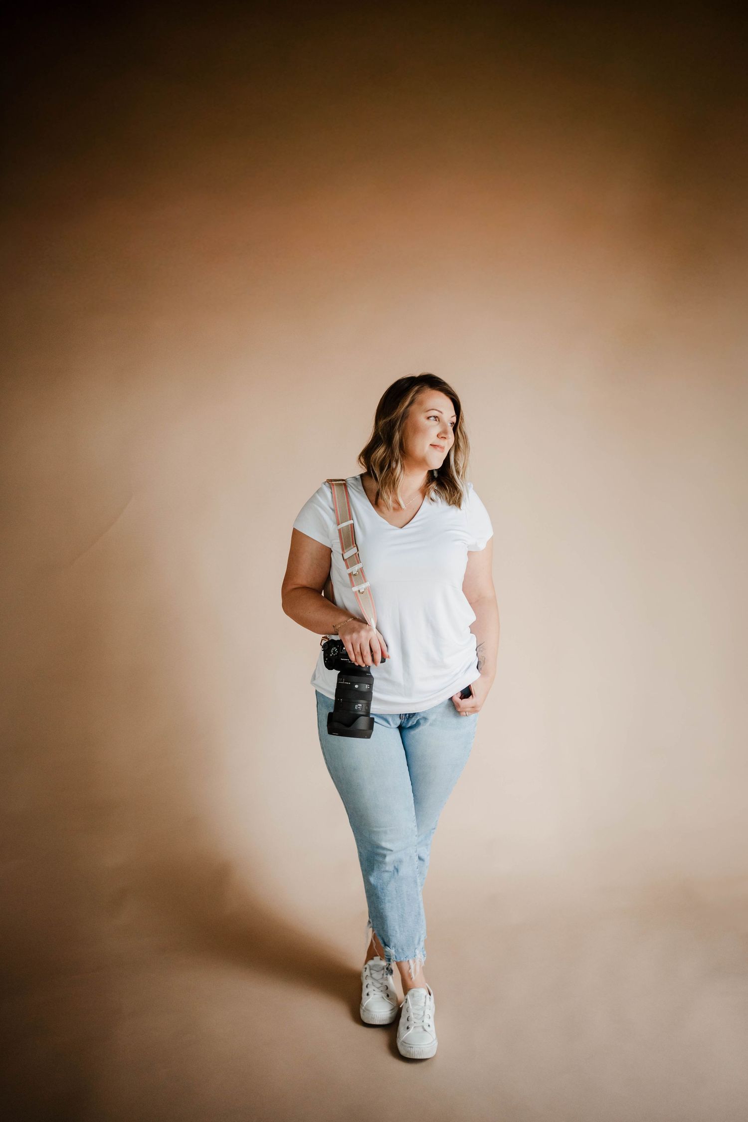 Female photographer in white tee and jeans holding camera with strap in a warm-toned studio setting.