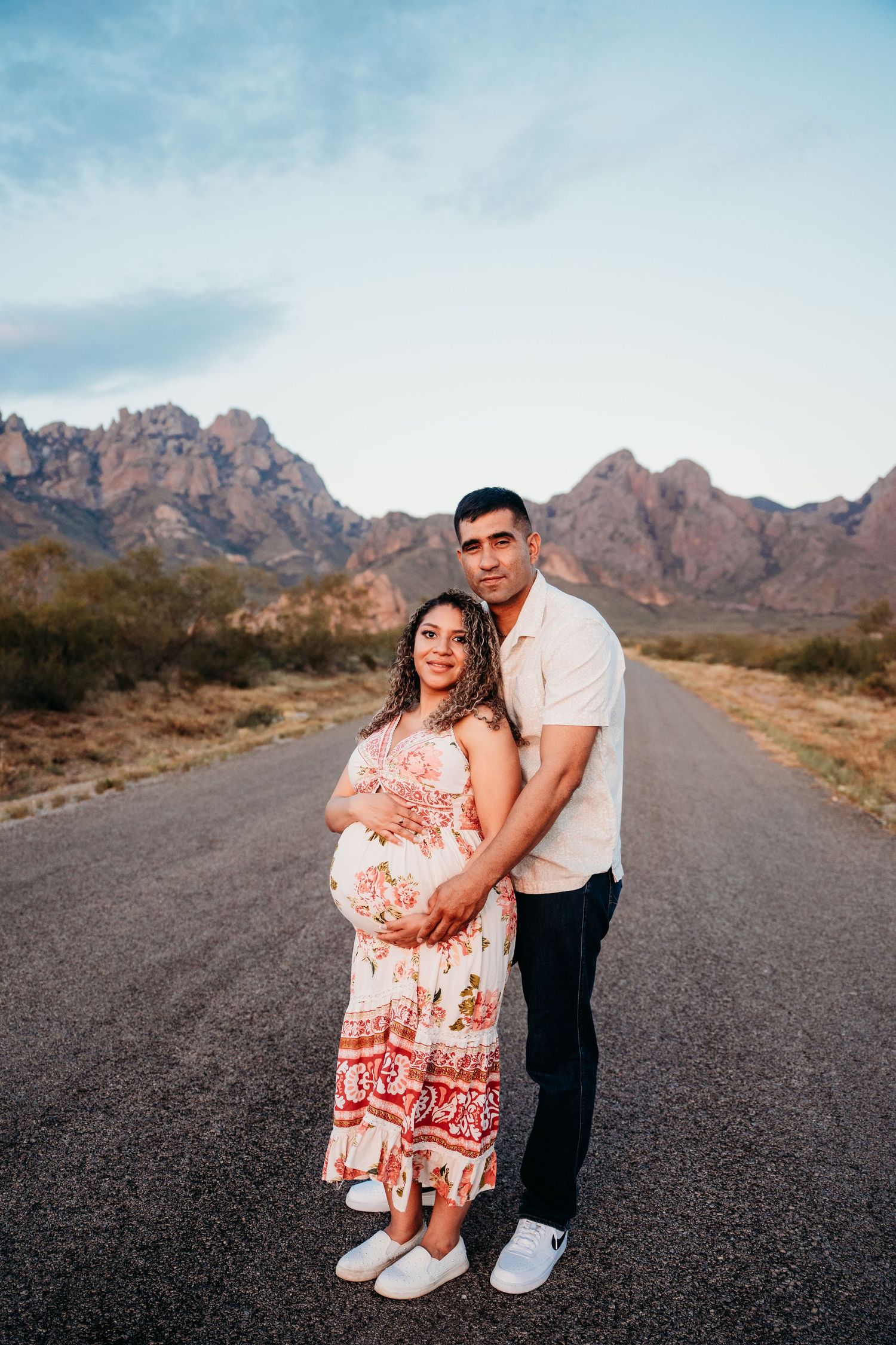 Pregnant couple embracing on open road with scenic mountain backdrop during maternity photo session.