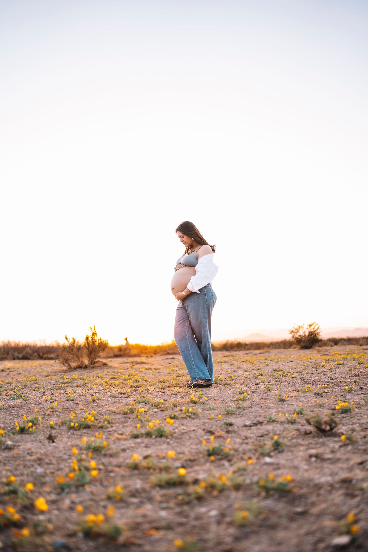Side profile of a pregnant woman holding her bump in an open desert landscape during a warm sunset.