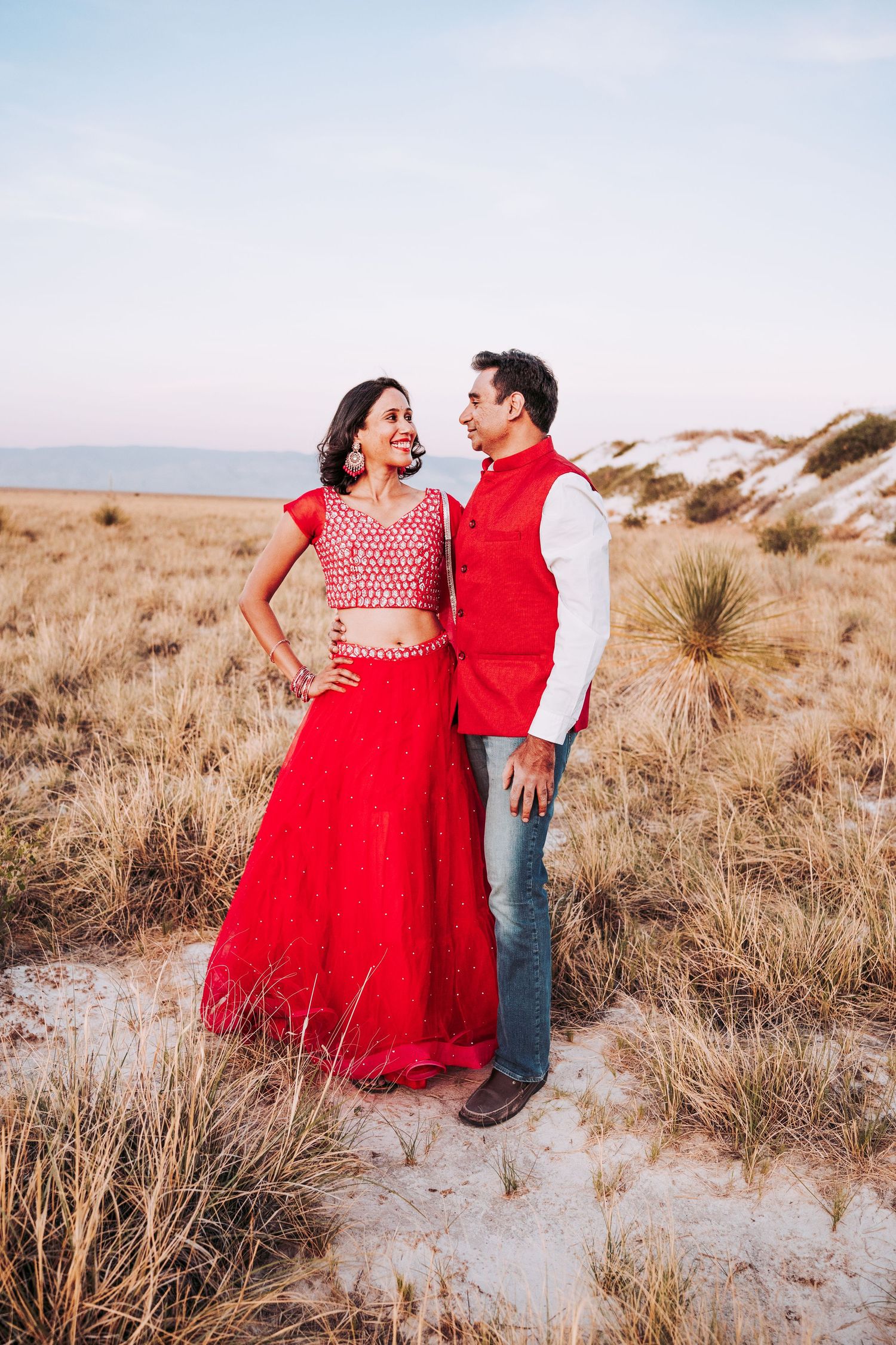 Indian couple in red traditional outfits standing together in a golden desert field.