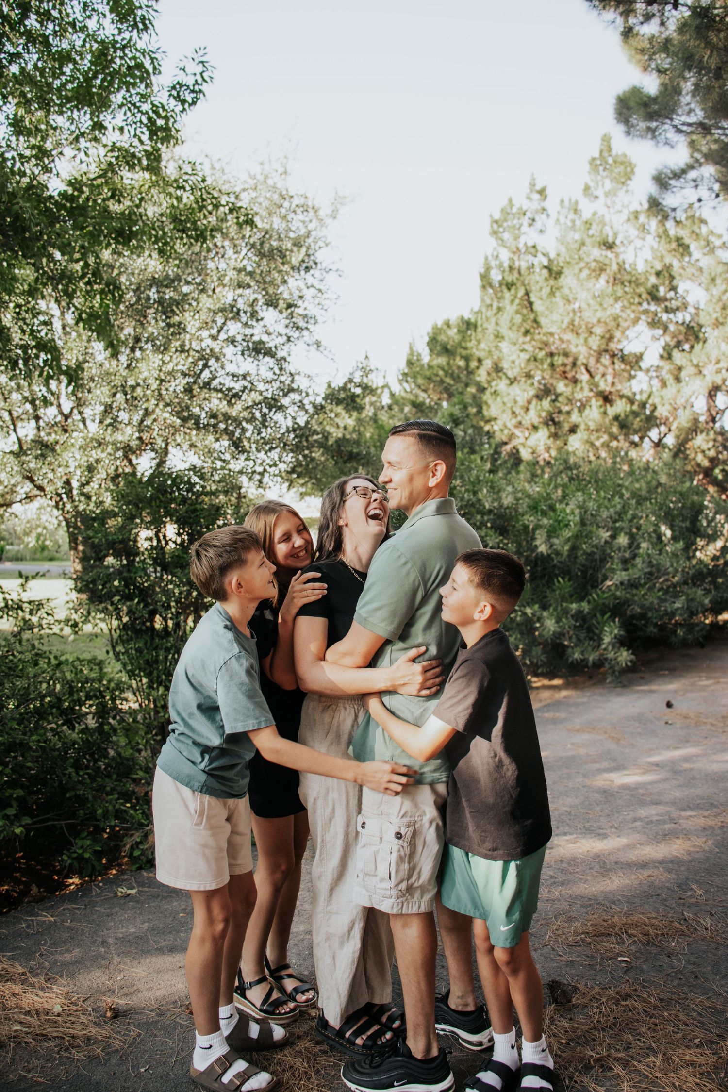 Family of five sharing a joyful group hug outdoors on a tree-lined path during a warm summer day.