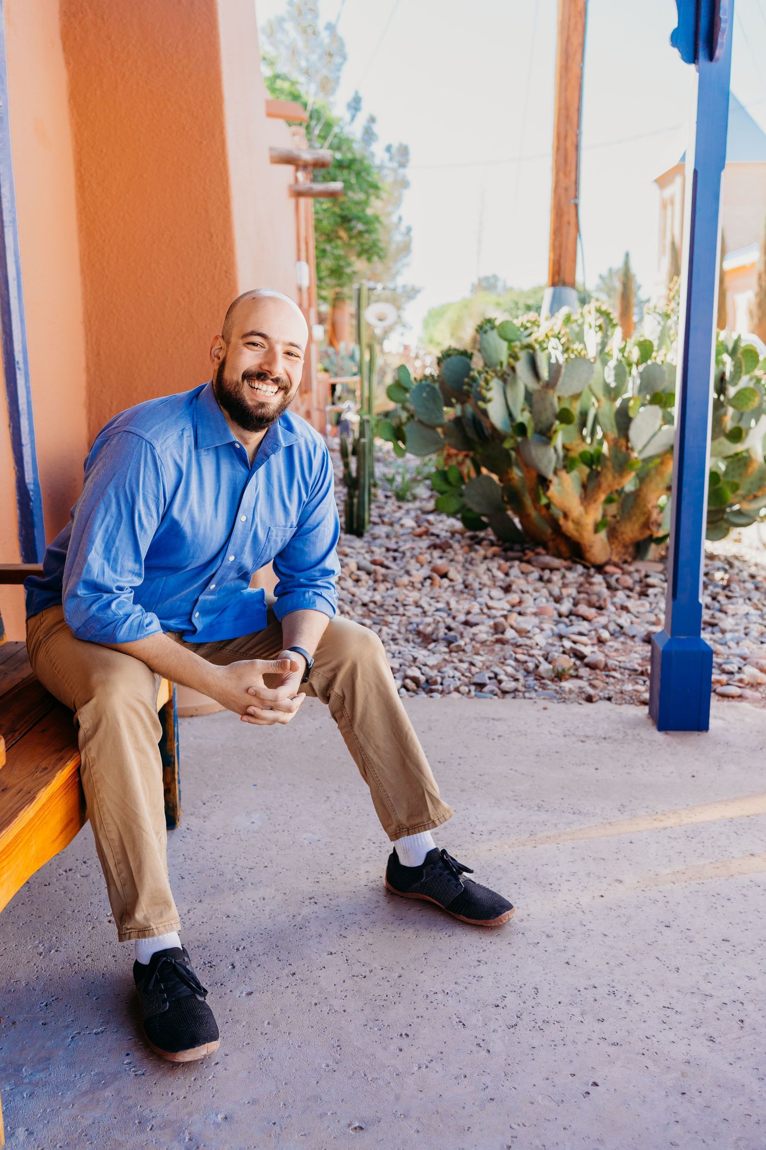 Smiling bald man in blue shirt seated on bench with desert landscape behind him.