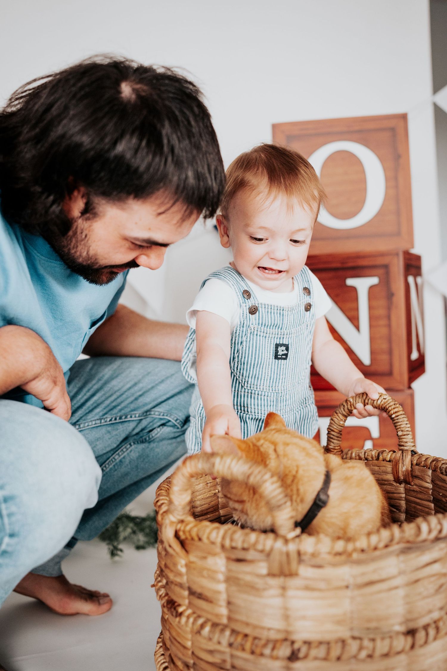 Father and baby boy smiling while exploring a wicker basket with an orange cat inside, with ONE blocks in background.