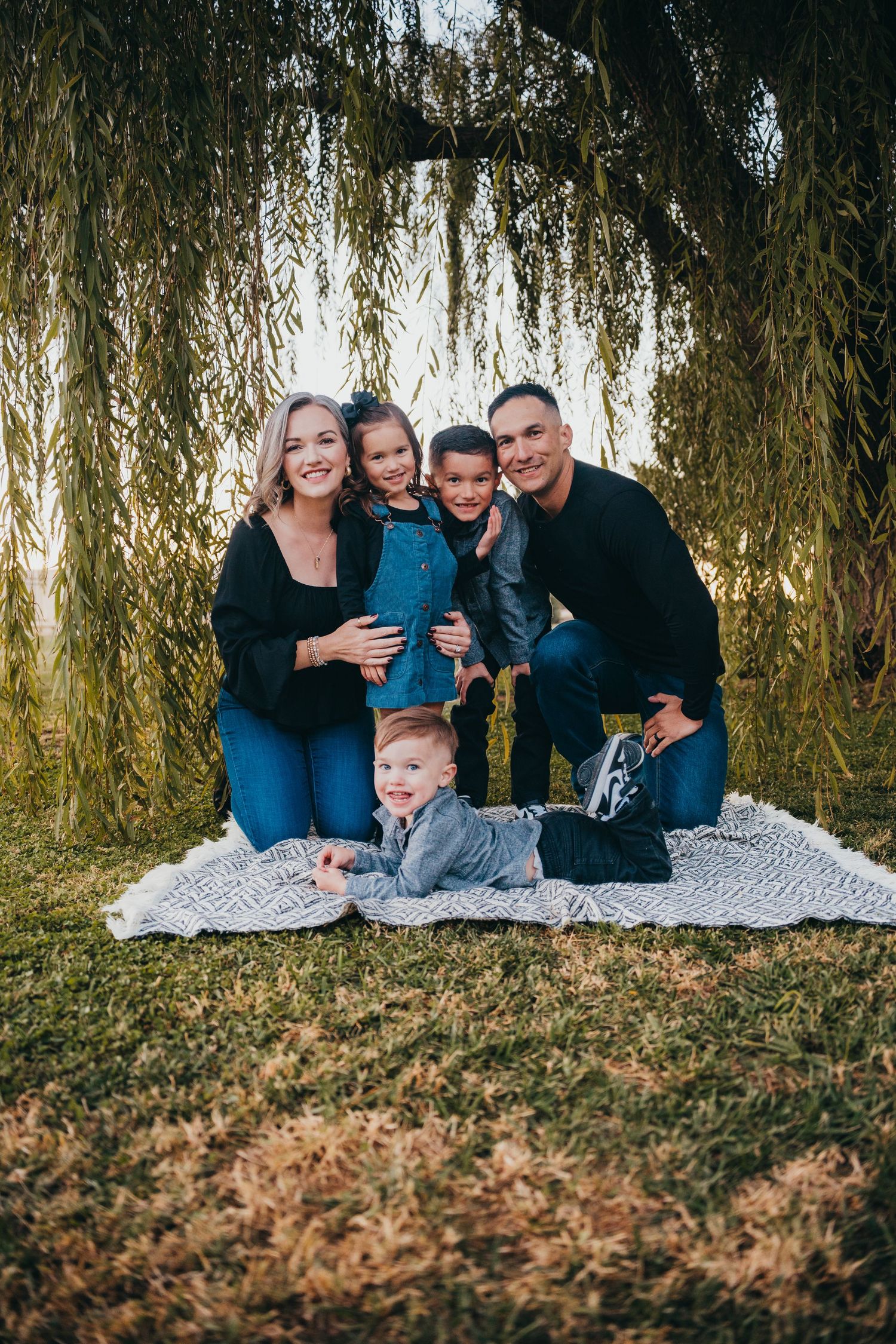 A happy family of five poses on a blanket under a weeping willow tree during an outdoor photo session.