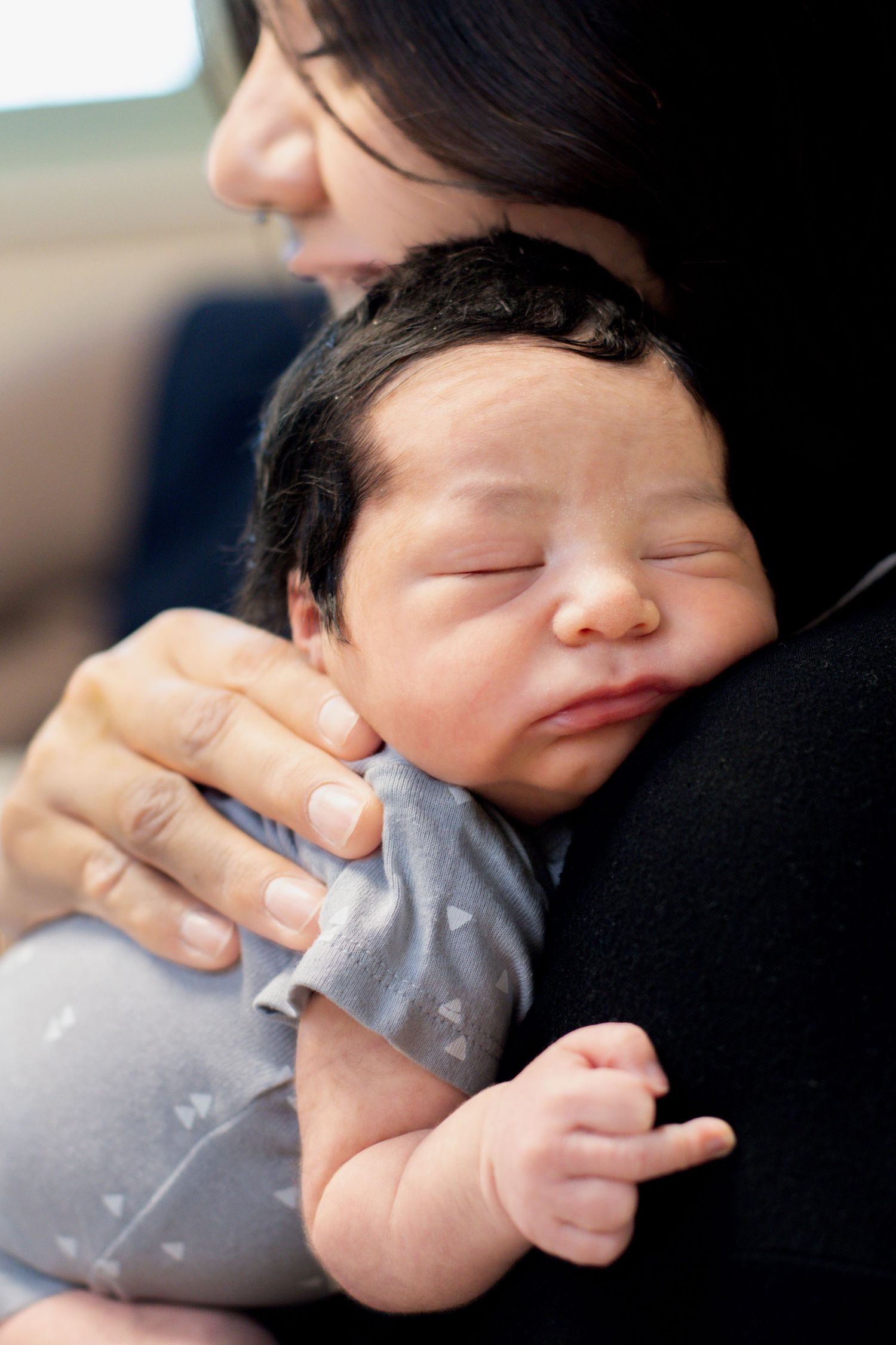 Mother cradling sleeping newborn baby with dark hair against her shoulder.