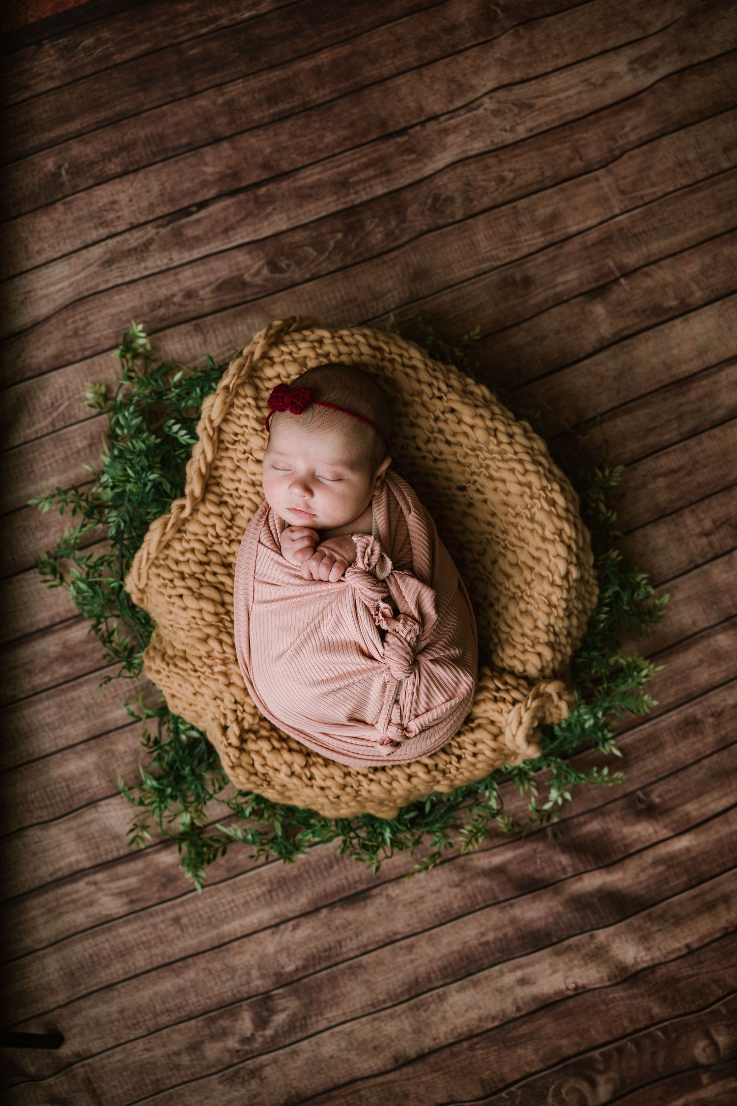 Overhead view of swaddled newborn in pink wrap resting in yellow knit bowl on wood.