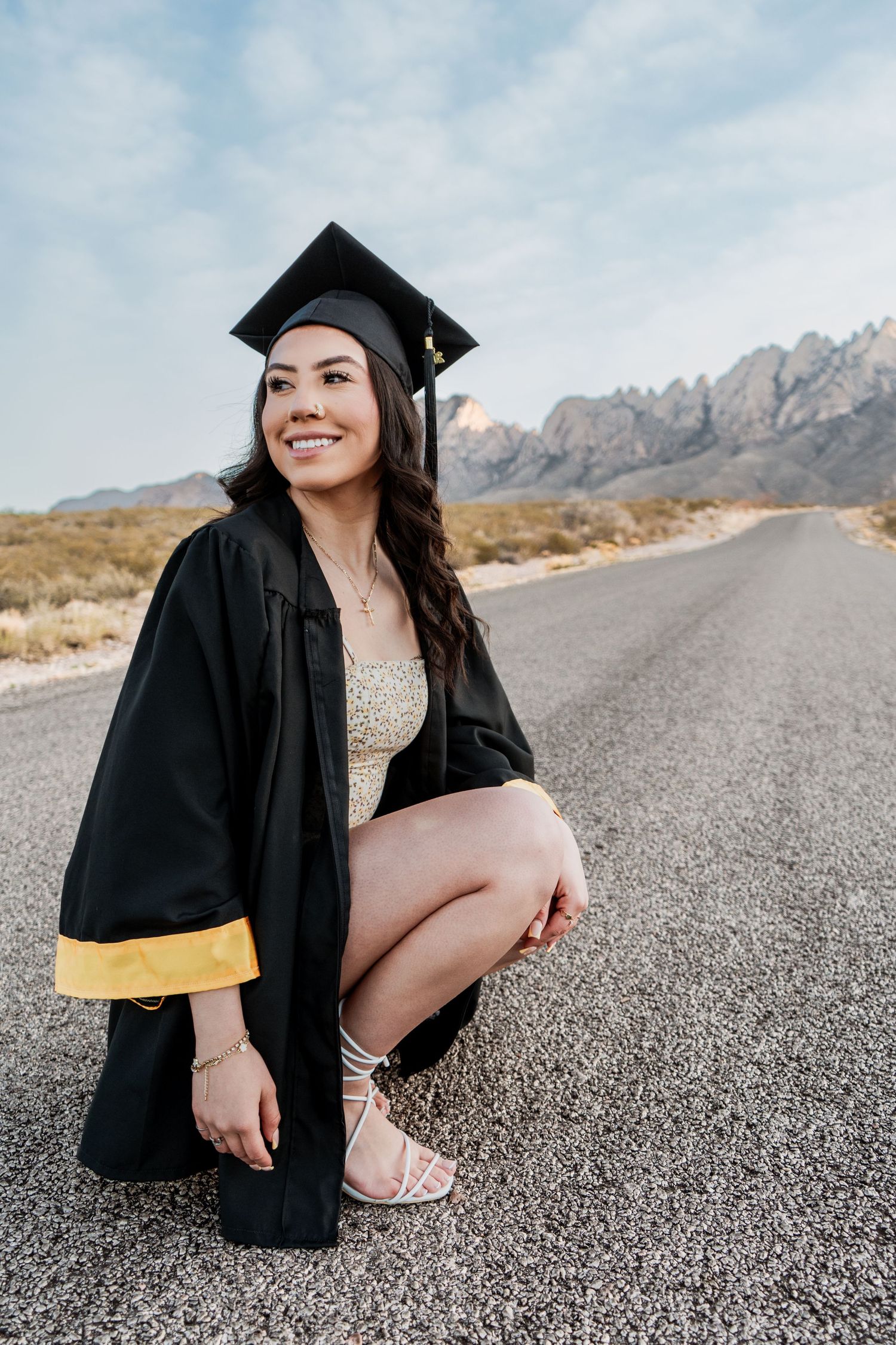 Happy female graduate in black mortarboard and gown poses crouching on an open road with mountains in background.