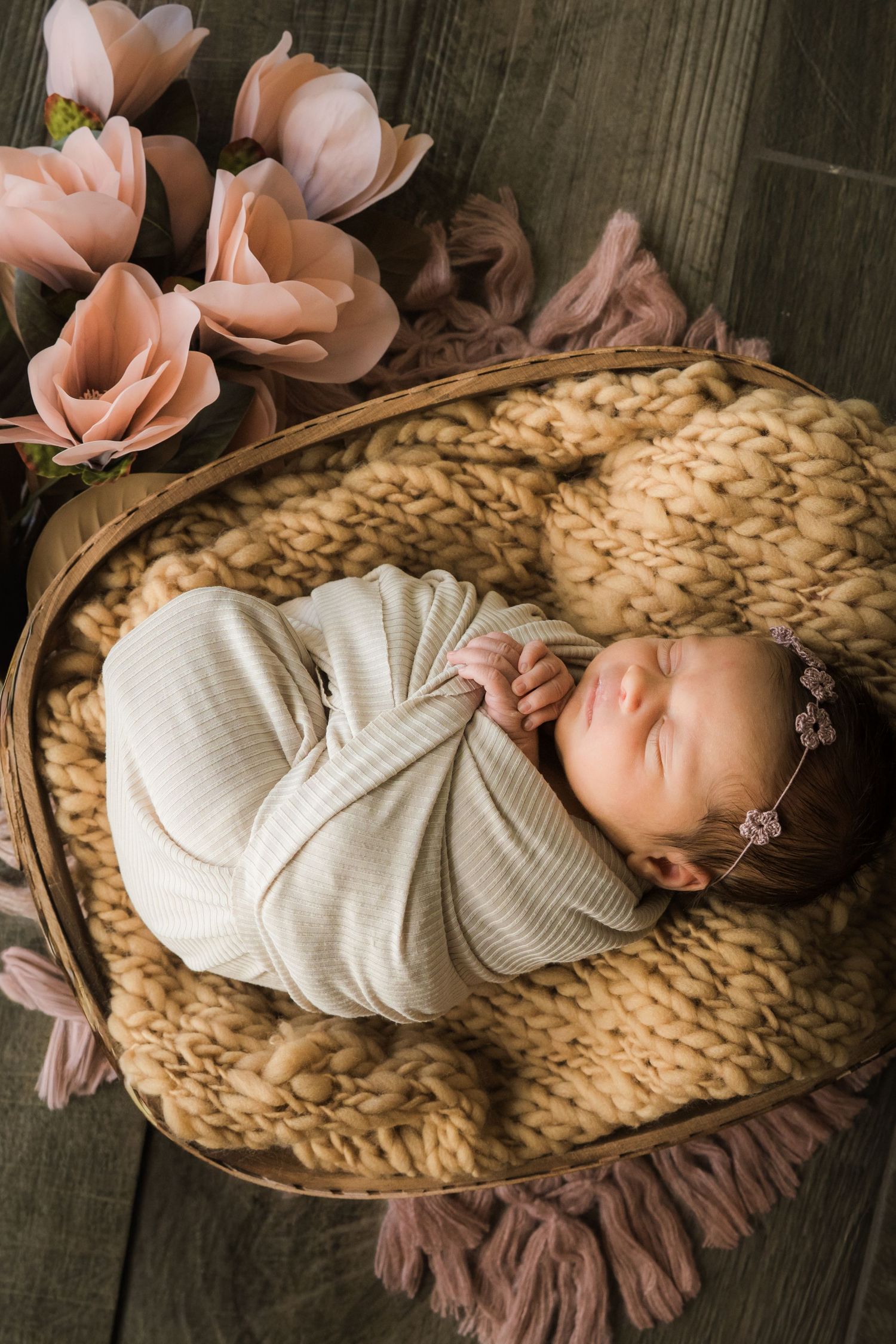 Newborn baby girl swaddled in striped wrap sleeping in a basket with pink flowers and knit blanket.