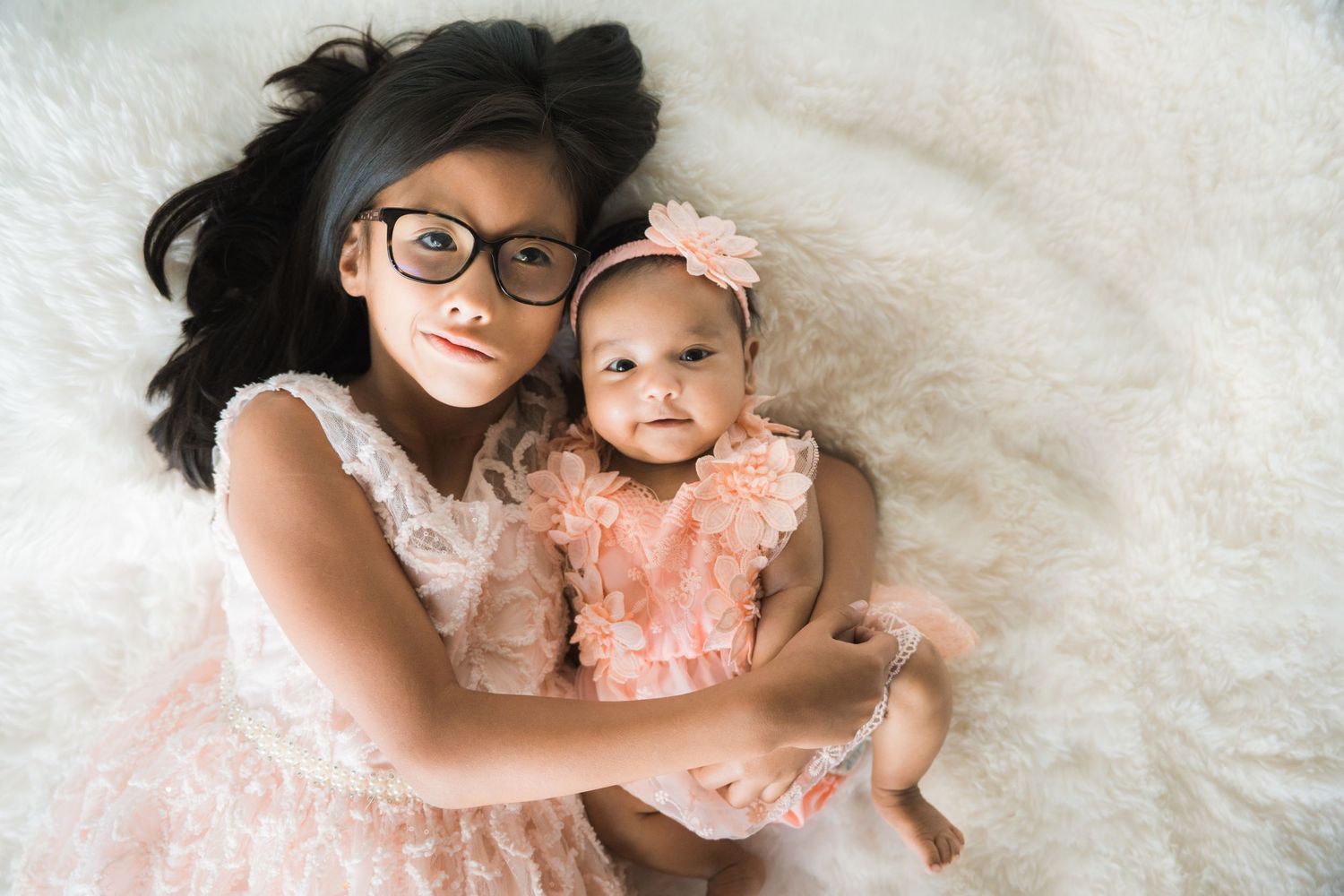 Young girl with glasses lying beside newborn baby sister in matching pink lace dresses.