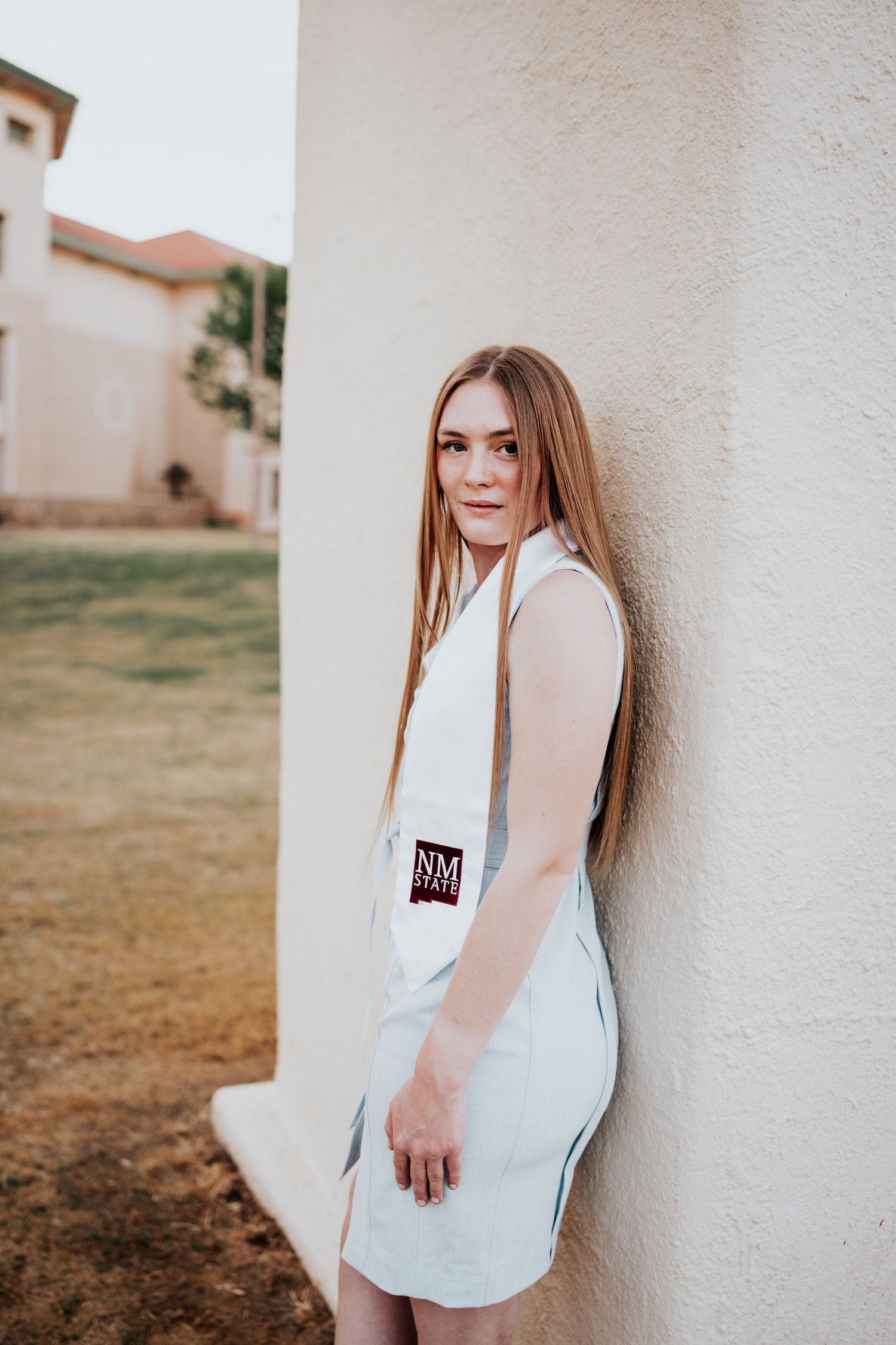 Blonde graduate in light blue dress with NM State stole leaning against white wall.