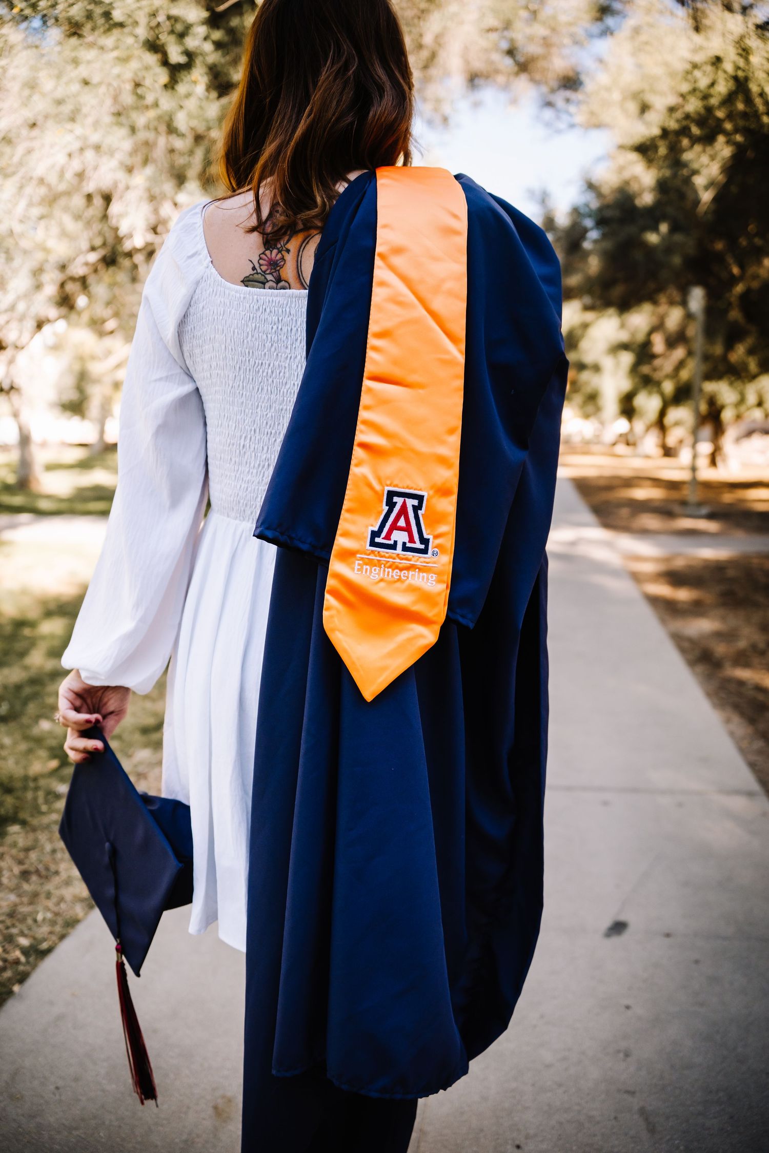 Back view of a female University of Arizona graduate in navy gown with orange stole holding cap.