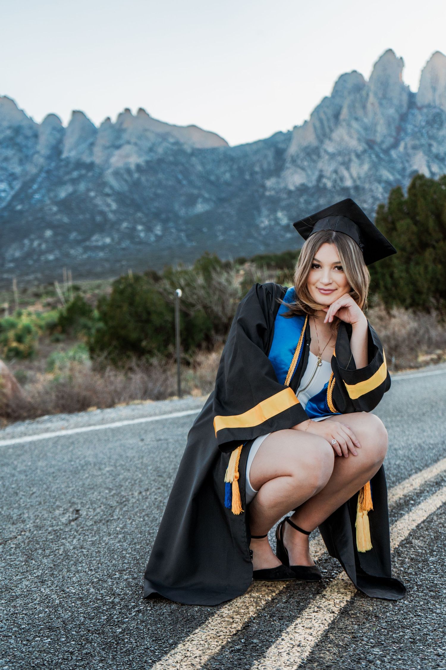 Smiling graduate in cap and gown crouching on road with rocky mountain landscape in background.