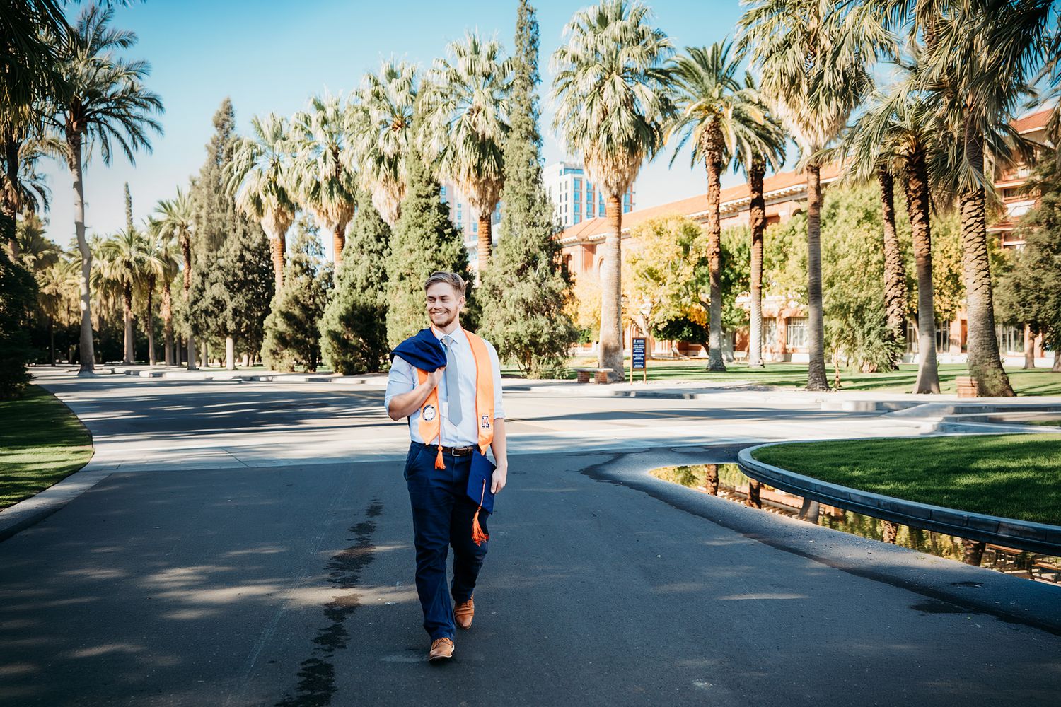 Graduate in cap and gown walks along a sunny palm tree-lined campus path, holding diploma and bag.