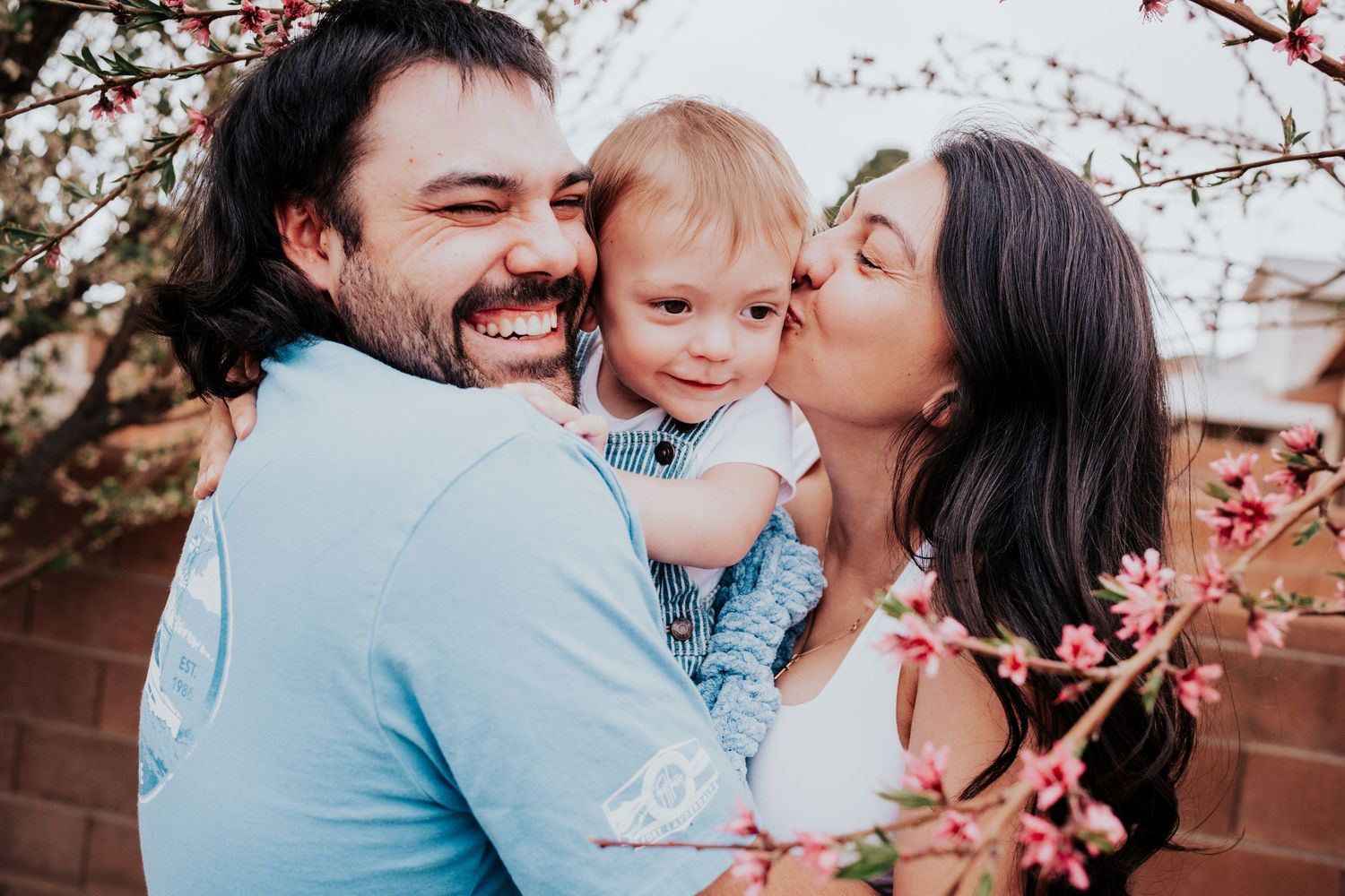 Mother kissing baby boy on cheek while father laughs joyfully among pink spring blossoms outdoors.