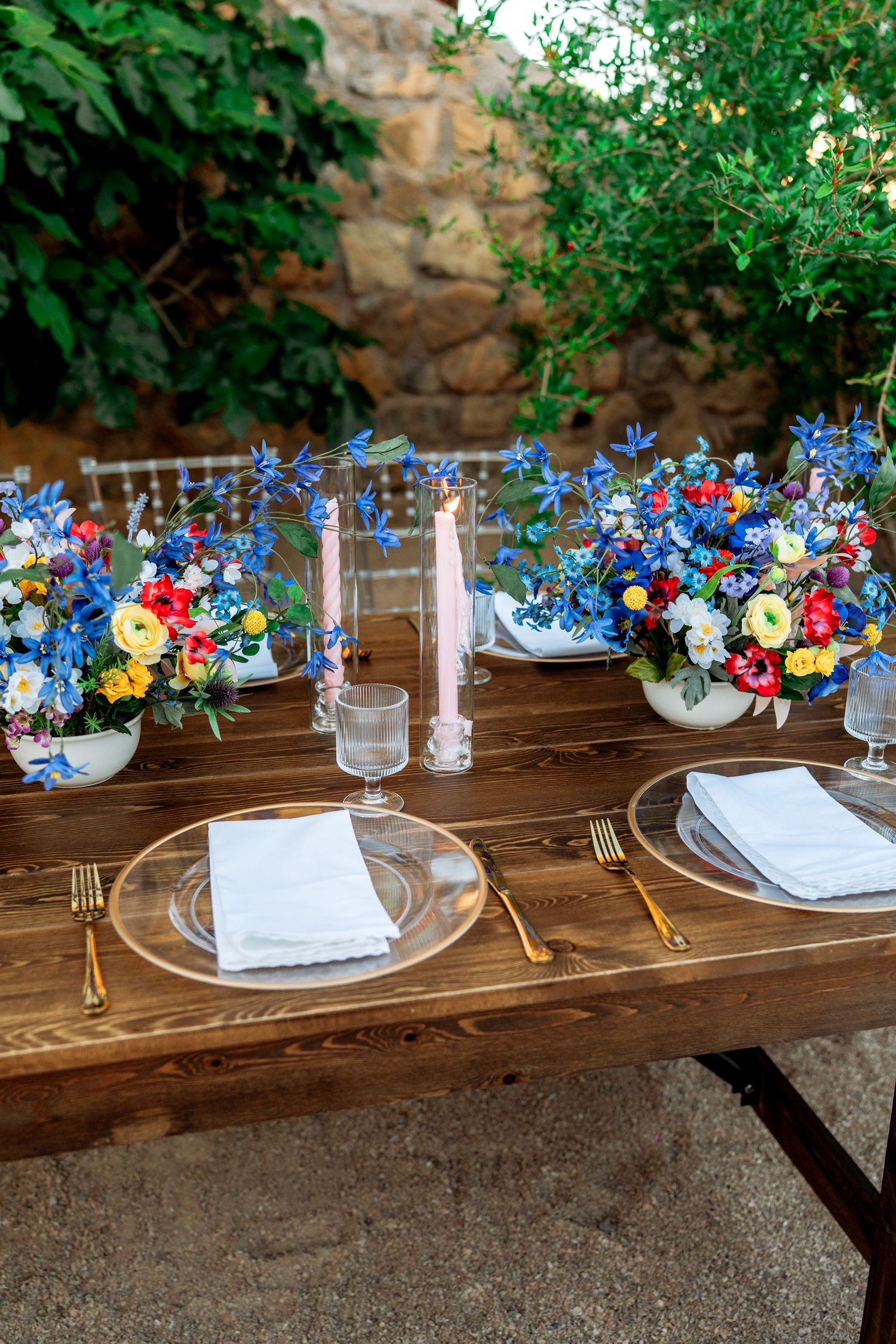 Rustic wooden table with gold cutlery, clear plates, pink candles, and vibrant blue and red floral centerpieces outdoors.