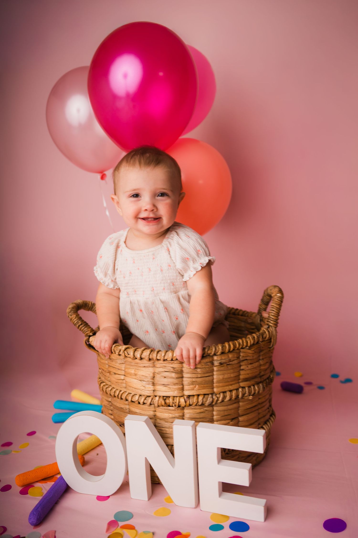 Smiling baby sitting in wicker basket with pink balloons and ONE letters for first birthday celebration.