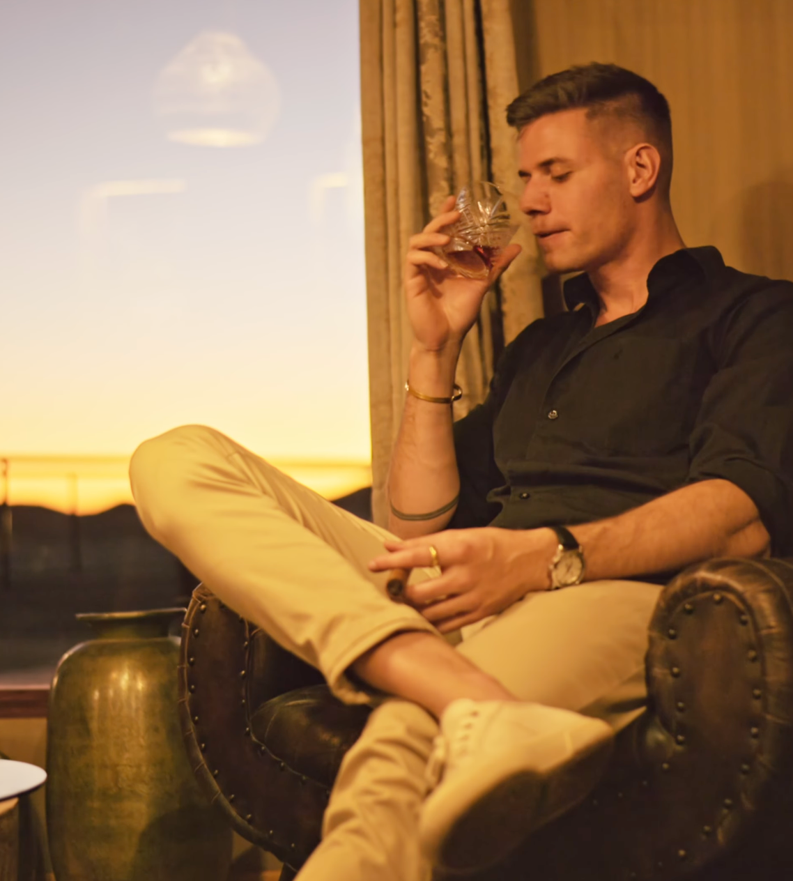 A stylish man in a black shirt and khaki pants relaxes in a leather chair, sipping whiskey at sunset.