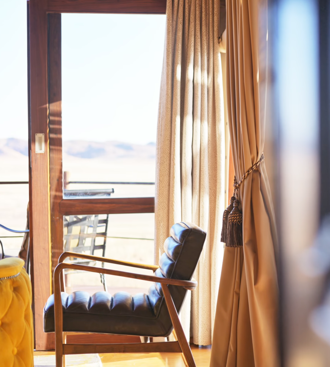 Elegant desert lodge room with leather chair, sheer curtains, and panoramic sand dune views through wooden framed windows.