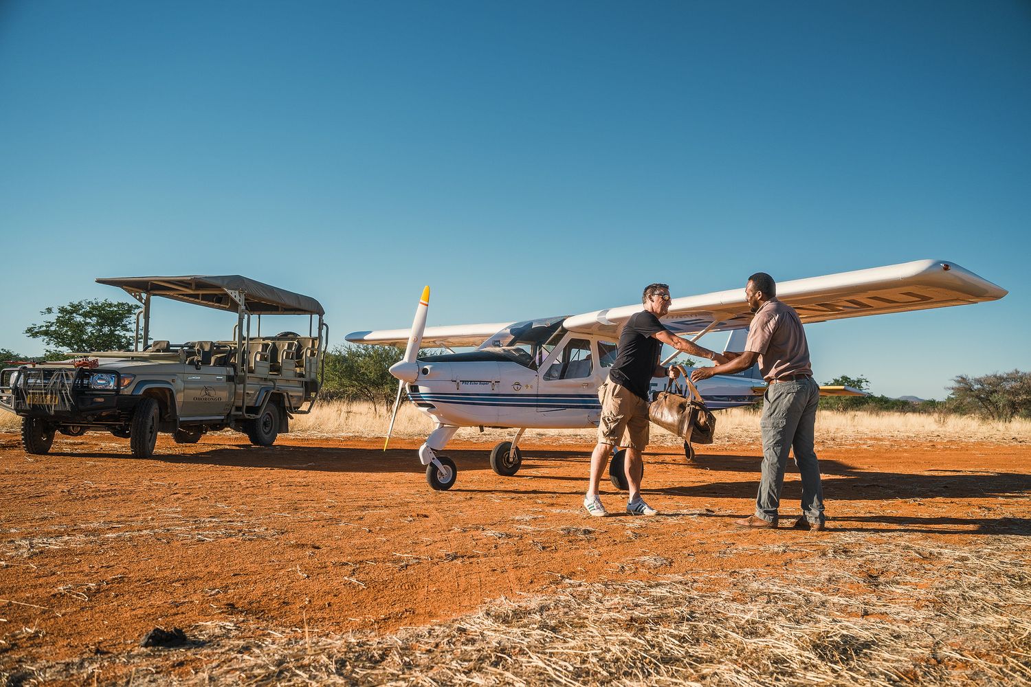 Two people shake hands near a small charter plane on a red dirt airstrip with a safari jeep parked nearby.