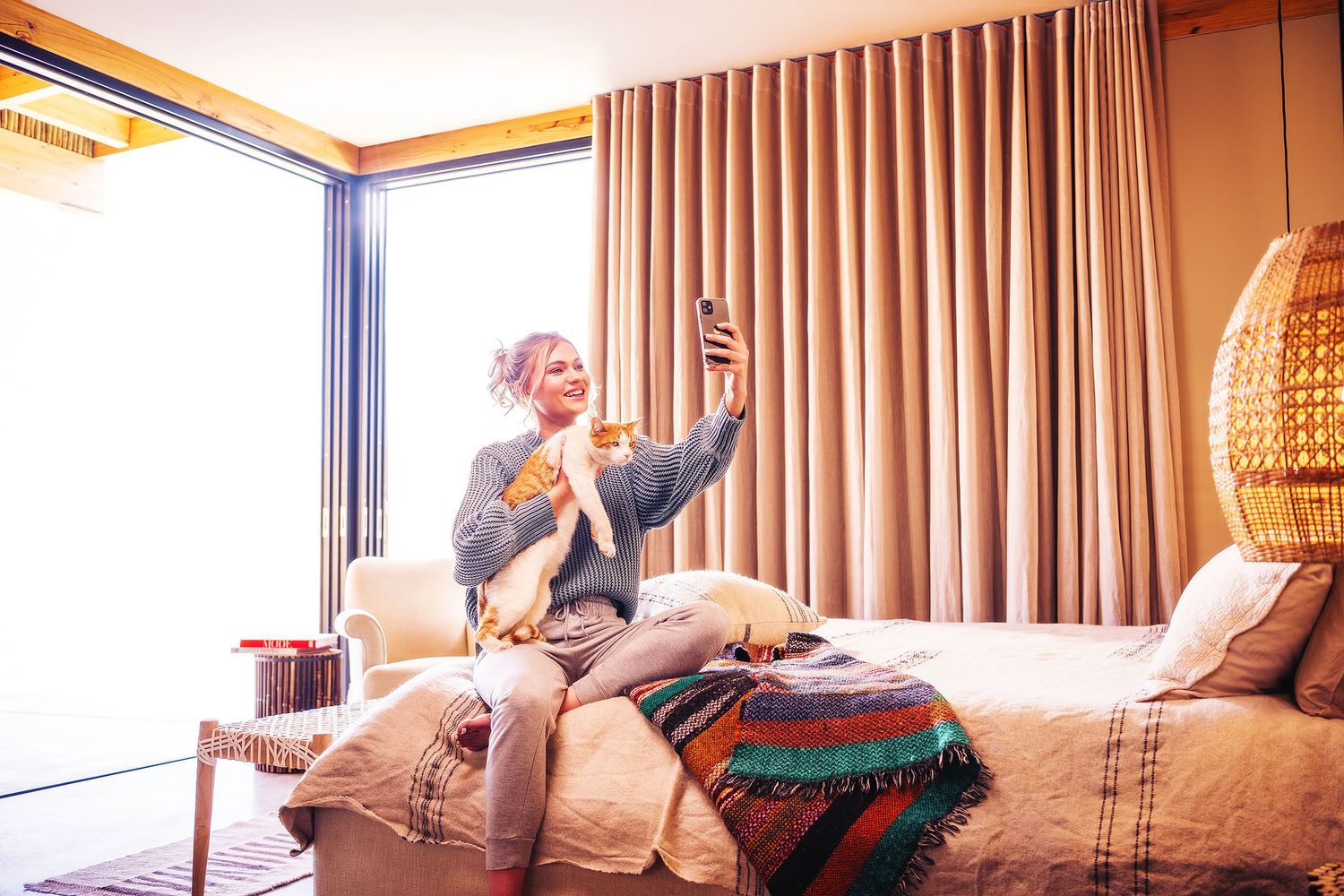 Woman taking a selfie with her dog on a cozy hotel bed near large windows with warm curtains.