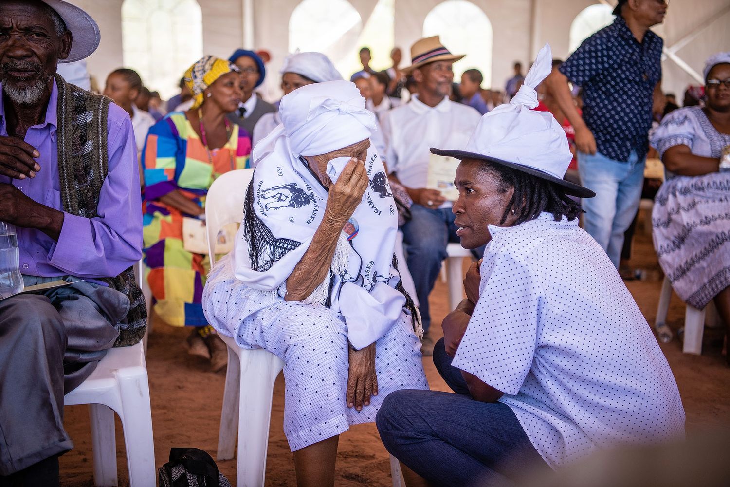 A woman in white traditional attire weeps while a man in a white hat consoles her at a cultural gathering.