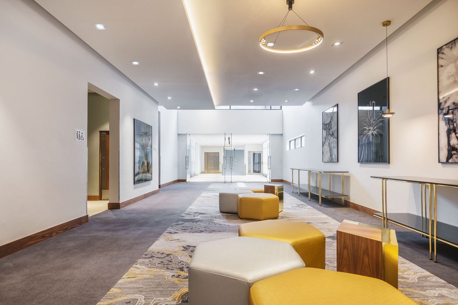 Modern hotel lobby with yellow hexagonal seating, gold pendant light, gray carpet, and bright white walls with artwork.