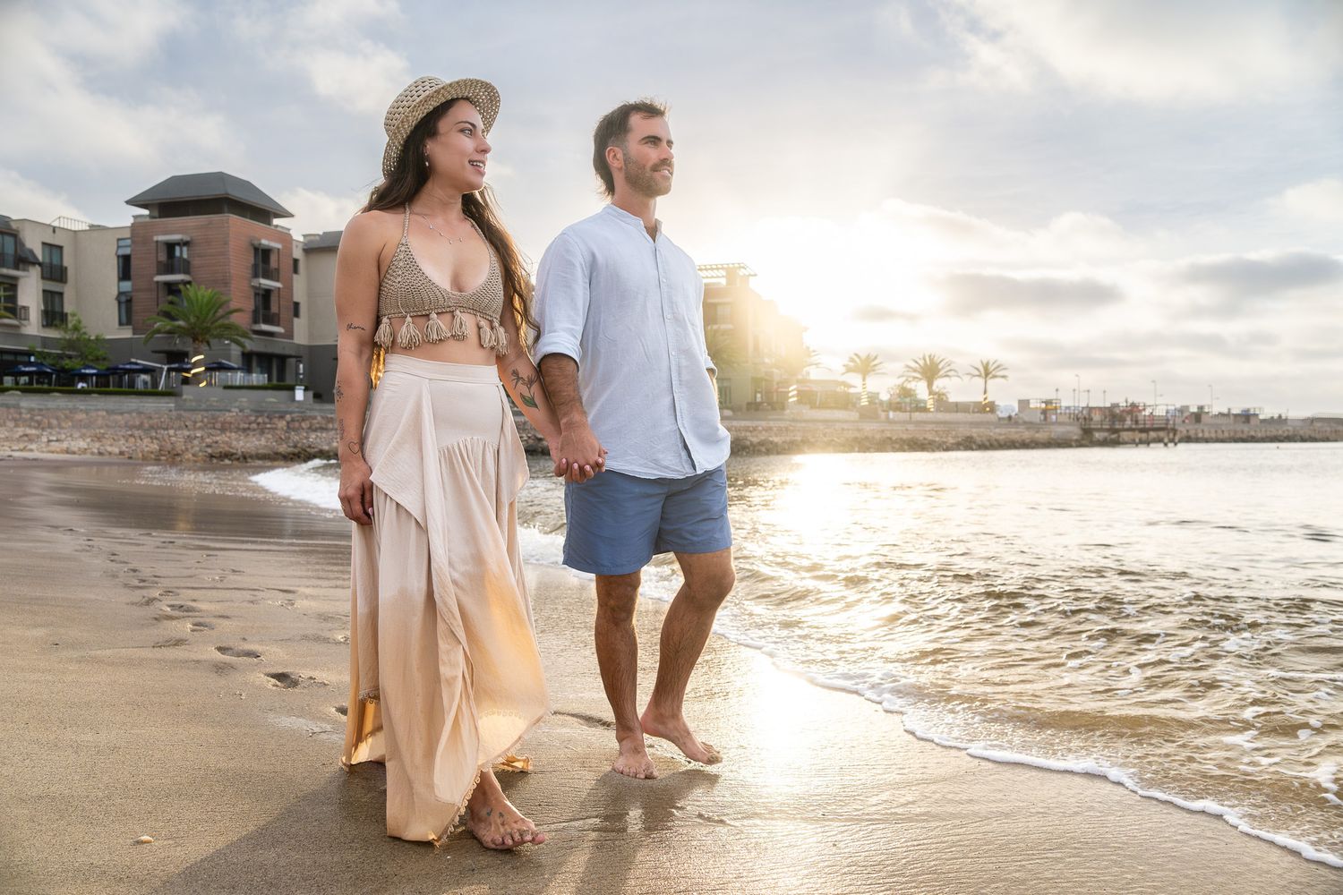 Couple holding hands walking barefoot on a sandy beach at golden sunset near a tropical resort.