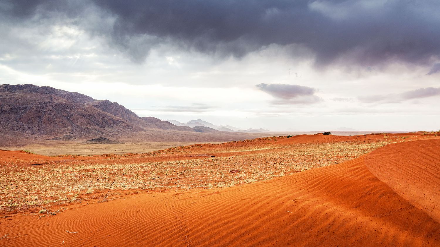 Vast orange sand dunes stretch across an arid desert landscape beneath dramatic storm clouds with mountains in the distance.