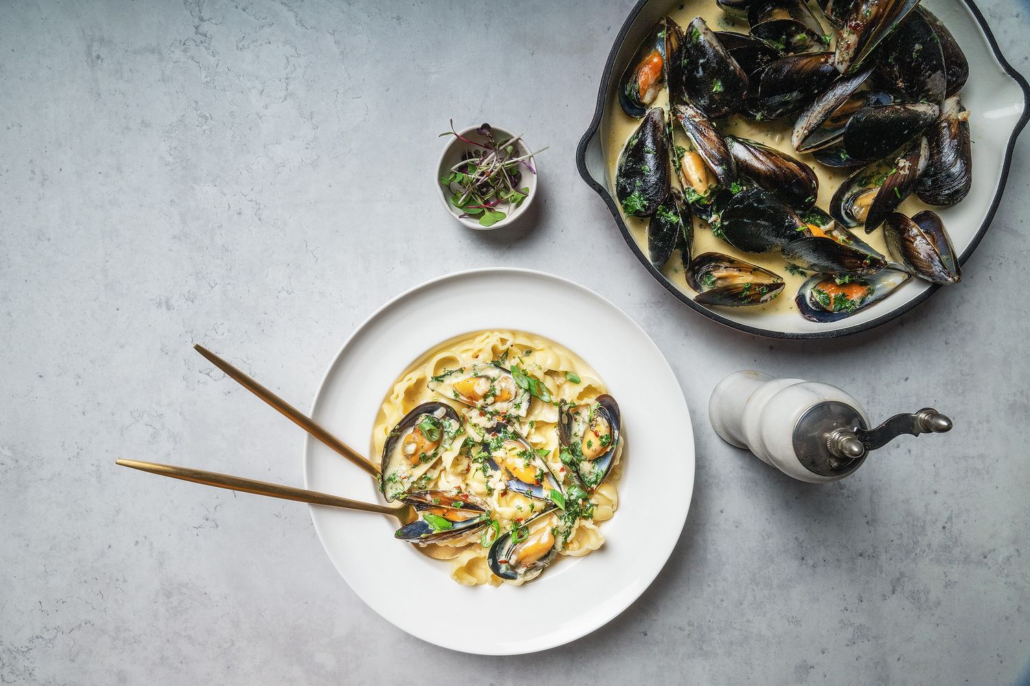 Steamed mussels in white wine broth served in a white bowl with chopsticks, alongside a pan of mussels on gray surface.
