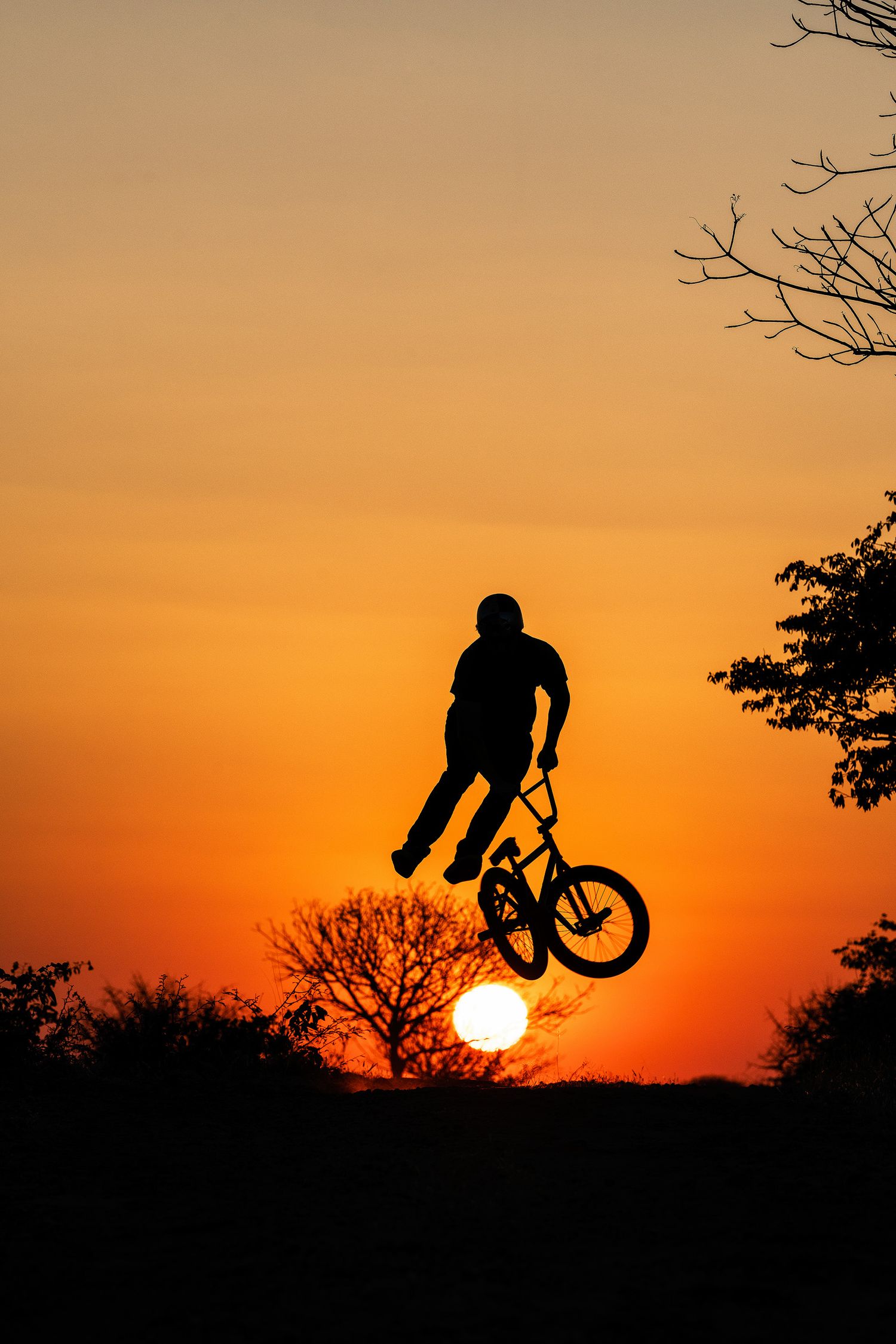 Silhouette of a BMX rider performing a jump trick against a vibrant orange sunset sky with trees in the background.