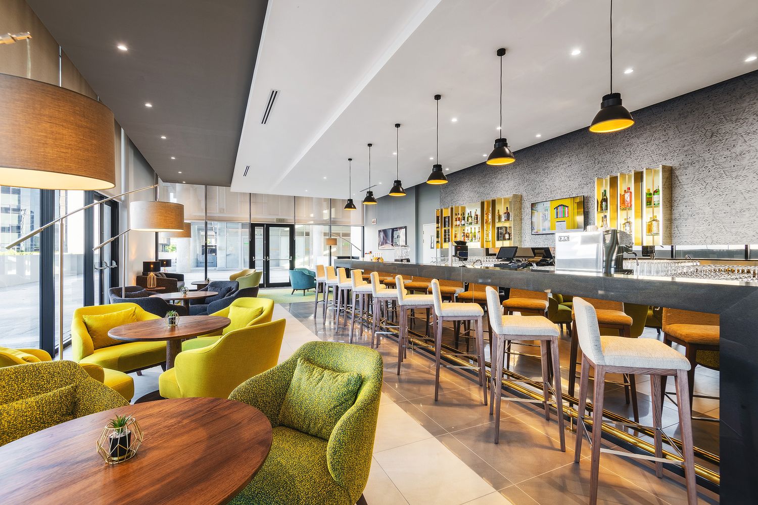 Modern hotel bar with yellow-green chairs, pendant lights, and a sleek counter with bar stools.