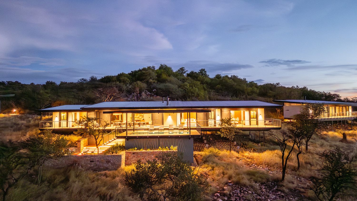 Illuminated luxury safari lodge glowing at dusk nestled among rocky African bushveld landscape.