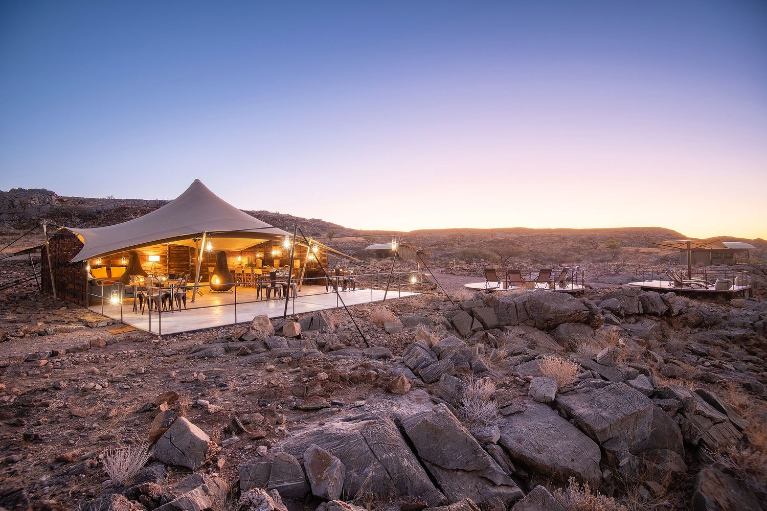 Luxury glamping tent illuminated at dusk in a rocky desert landscape with panoramic views.