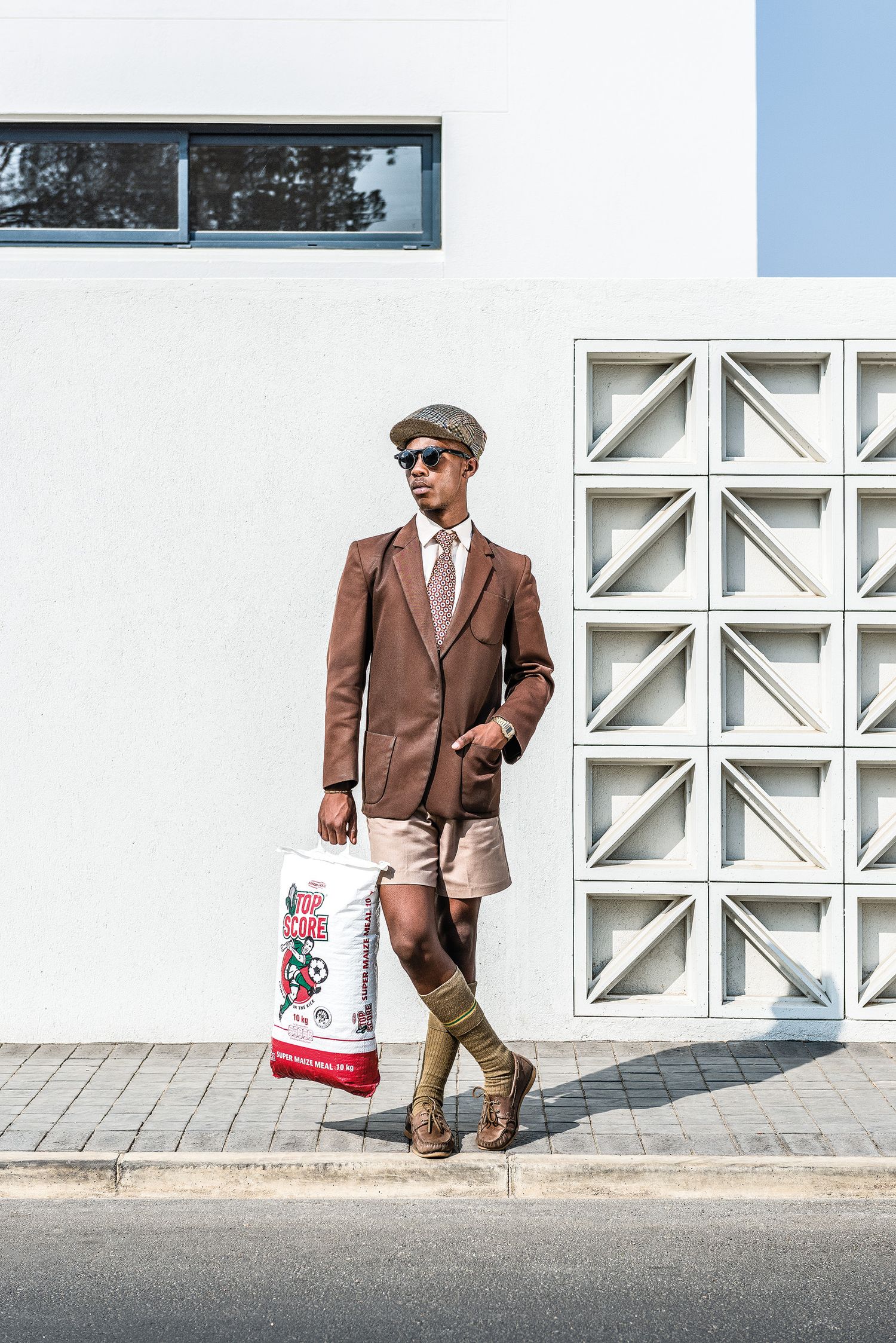 Stylish man in brown blazer, shorts, knee socks and flat cap posing against a white urban wall.