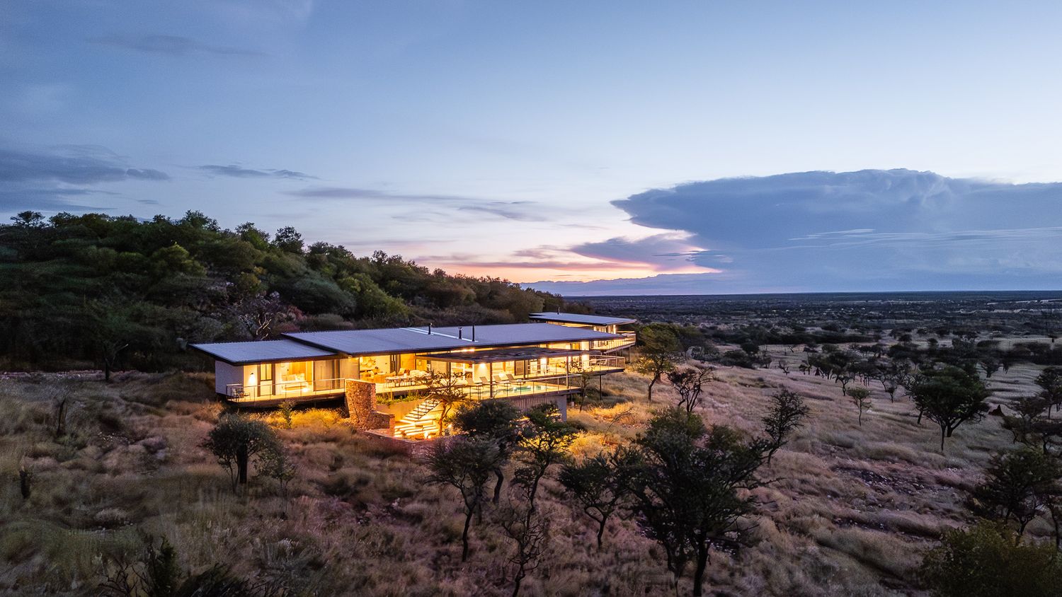 Illuminated modern safari lodge glowing at dusk with panoramic views of African bushveld landscape.
