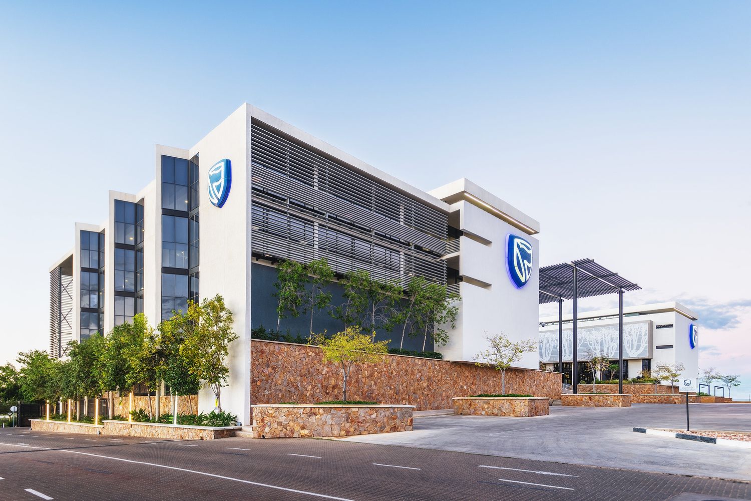 Modern multi-story commercial building with blue corporate logo, solar panels and landscaped entrance at dusk.