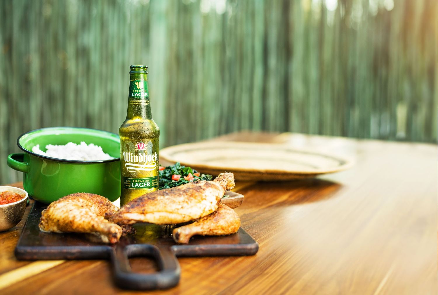 Grilled chicken pieces on a wooden board with a green beer bottle, bowl of rice, and dipping sauce outdoors.
