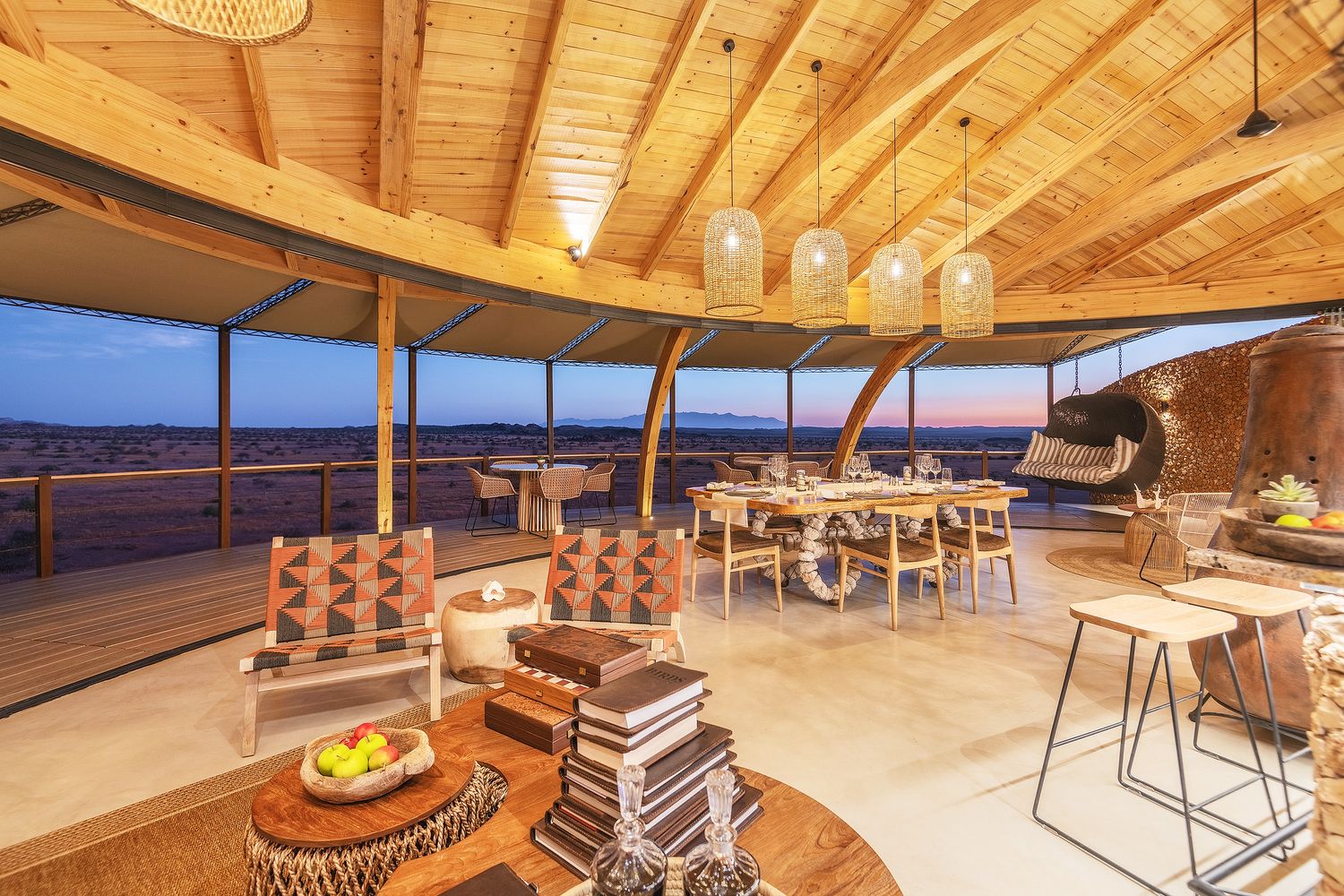 Elegant open-air safari lodge lounge with wooden curved ceiling, rattan furniture, and panoramic desert views at dusk.